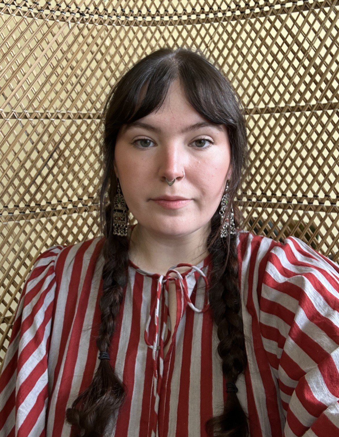 A young woman with dark brown hair styled in two braids, wearing large earrings and a black septum ring, dressed in a red and beige striped blouse, standing in front of a woven bamboo wall.