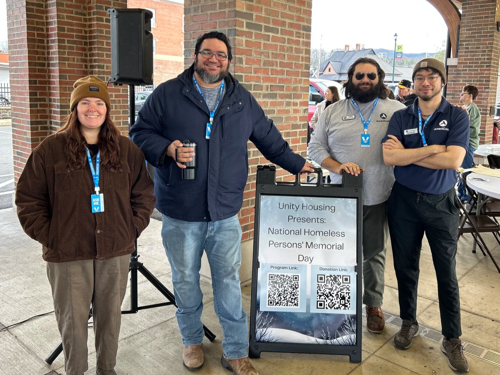 Group of four people standing outdoors with a sign for Unity Housing and a memorial event. The sign displays QR codes for program and donation links. The group includes a woman in a brown jacket and beanie, and three men, one with sunglasses and a gray shirt, and two in navy shirts. They are smiling and wearing lanyards.