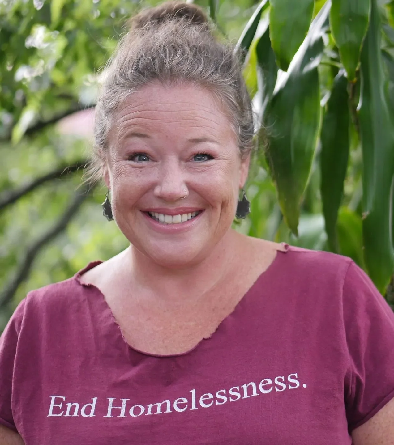 A woman with gray hair tied back, smiling, wearing a maroon shirt that says "End Homelessness." standing outdoors among green plants.