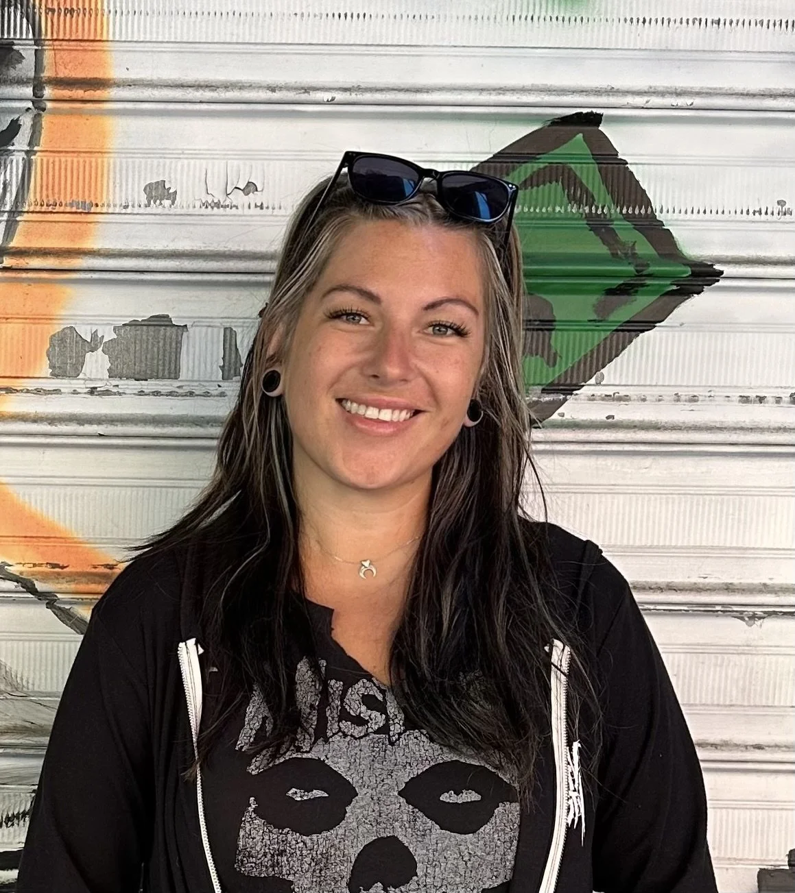 A woman with long dark hair, wearing a black hoodie over a black Zombie Skull graphic T-shirt, standing in front of a painted metal garage door.