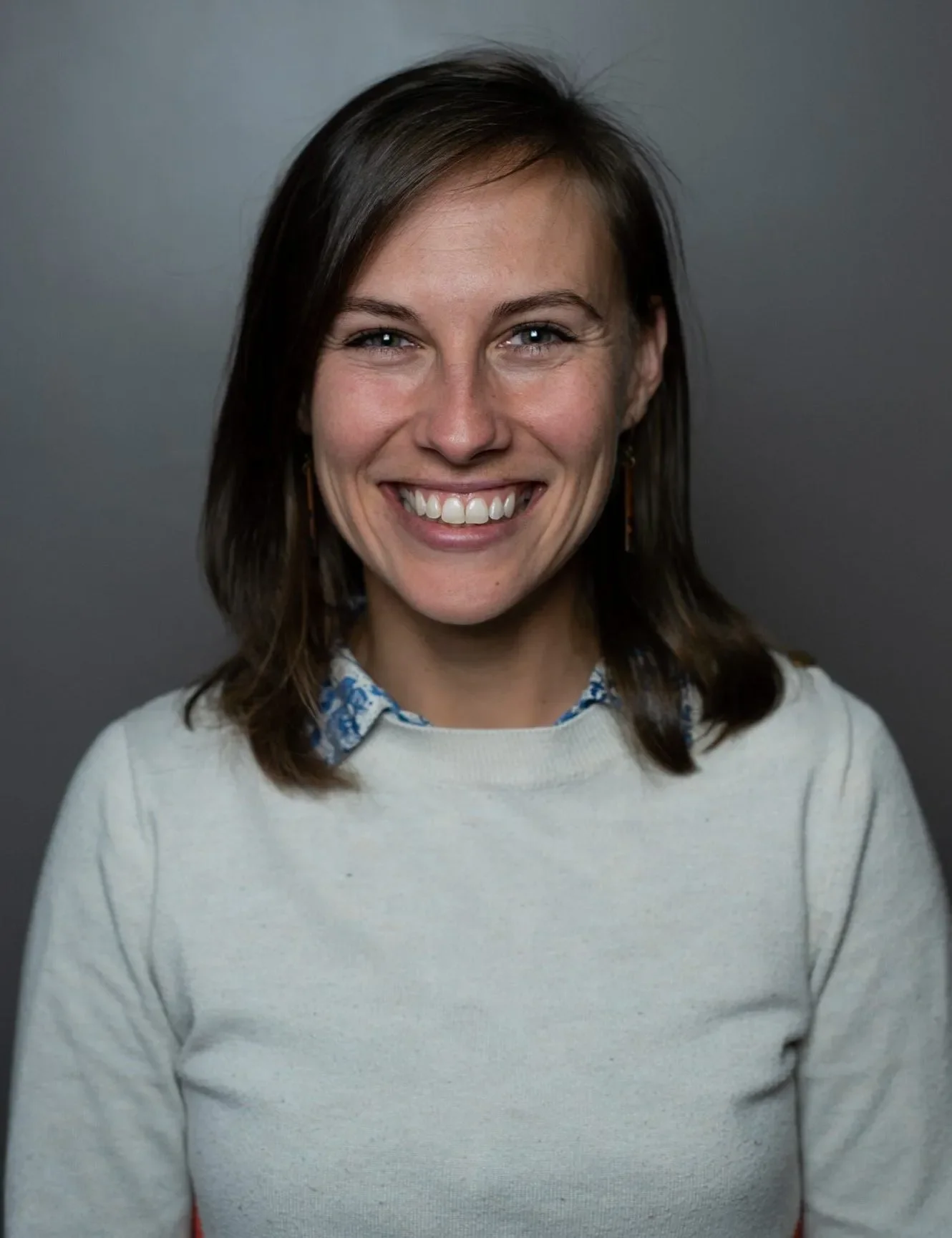 A woman with shoulder-length brown hair, blue eyes, and a bright smile, wearing a cream sweater with a collared shirt underneath, standing against a plain dark background.