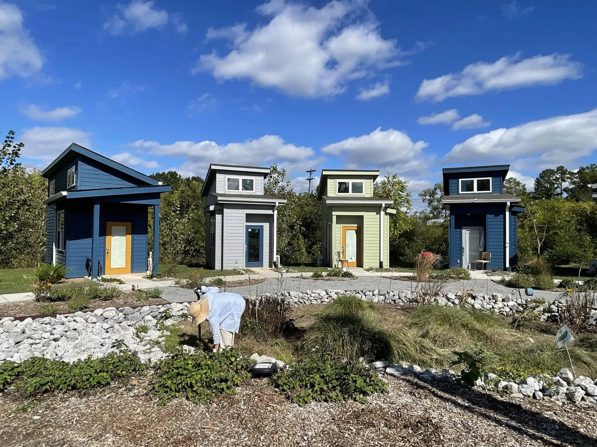 Colorful tiny houses with sloped roofs lined up in a garden area with plants and rocks, under a partly cloudy blue sky.