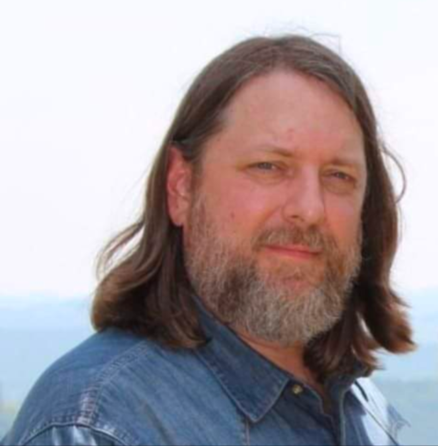 Portrait of a man with long brown hair and a beard, wearing a denim shirt, outdoors with a cloudy sky in the background.