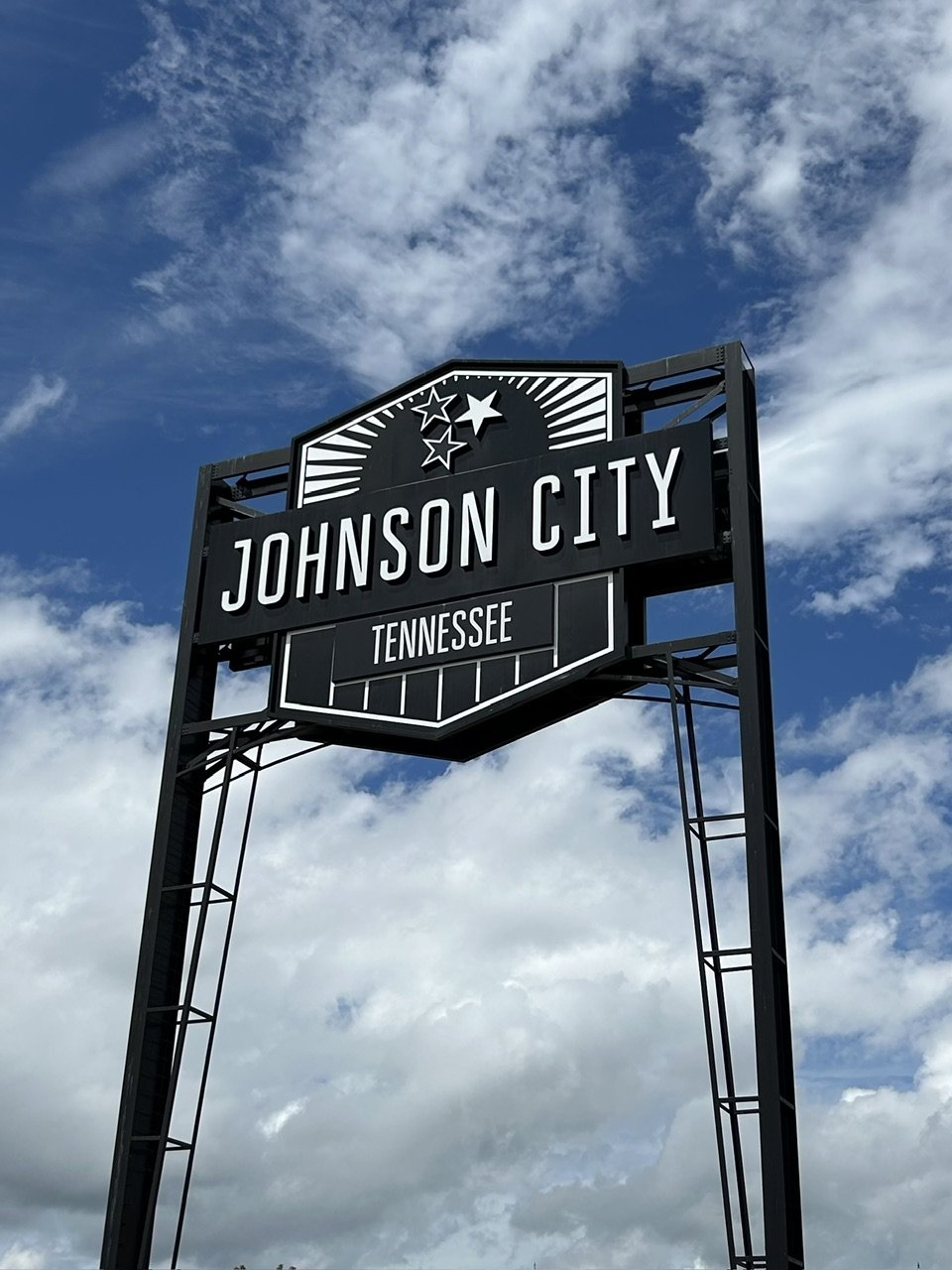 Black sign with white lettering reads 'Johnson City' and 'Tennessee', with stars and rays design, against a cloudy sky.