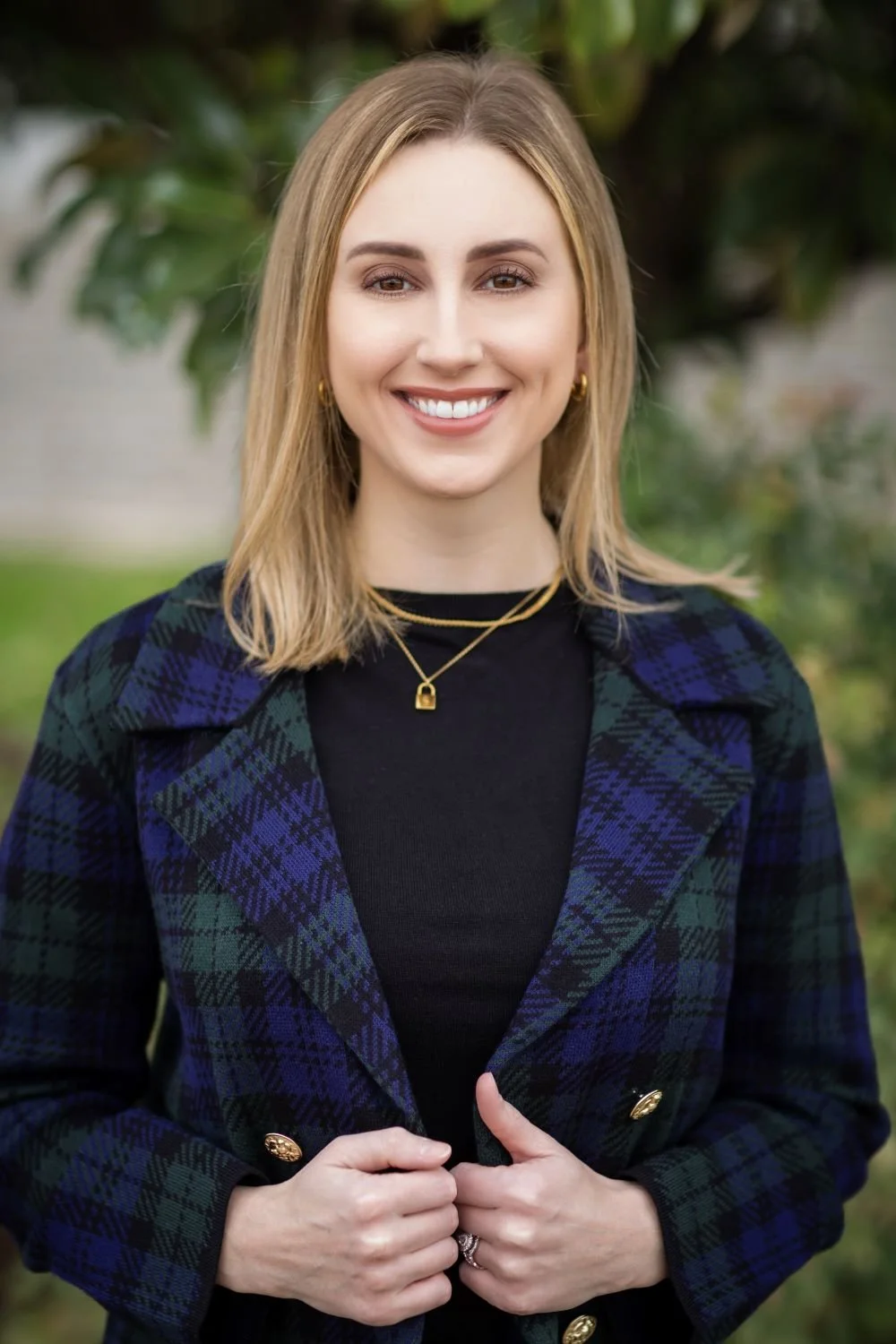 A woman with blonde hair smiling outdoors wearing a black top and a blue-green plaid blazer, accessorized with gold jewelry, standing in front of green foliage.