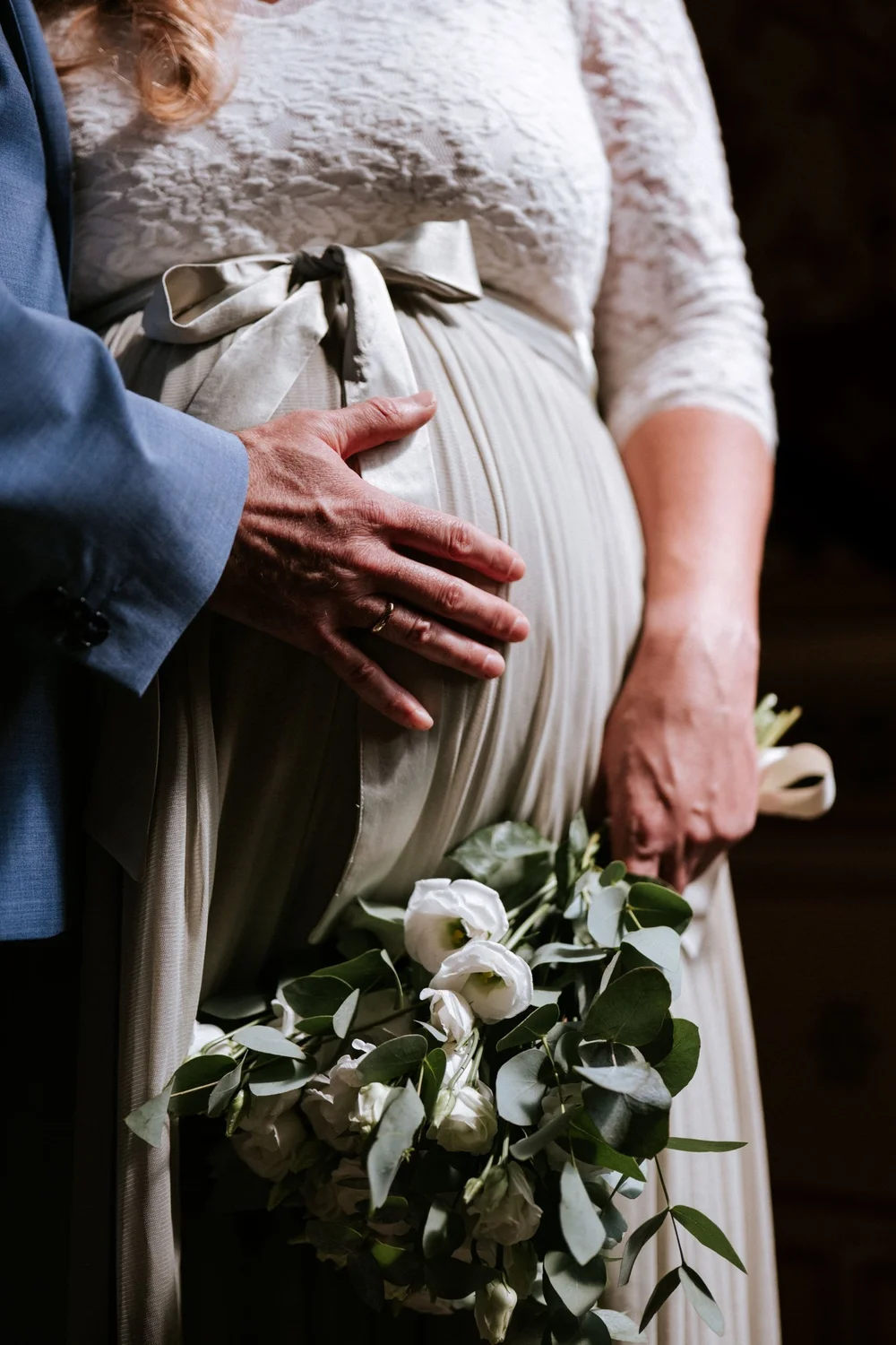 Schwangere Braut im Spitzenkleid mit Schleife und Brautstrauß, Hand des Bräutigams am Babybauch, Hochzeit in Korneuburg