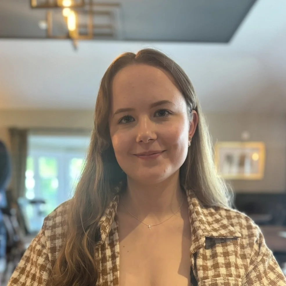 A woman with long wavy blonde hair, wearing a brown checkered jacket and a delicate necklace, smiling in an indoor setting with natural light.