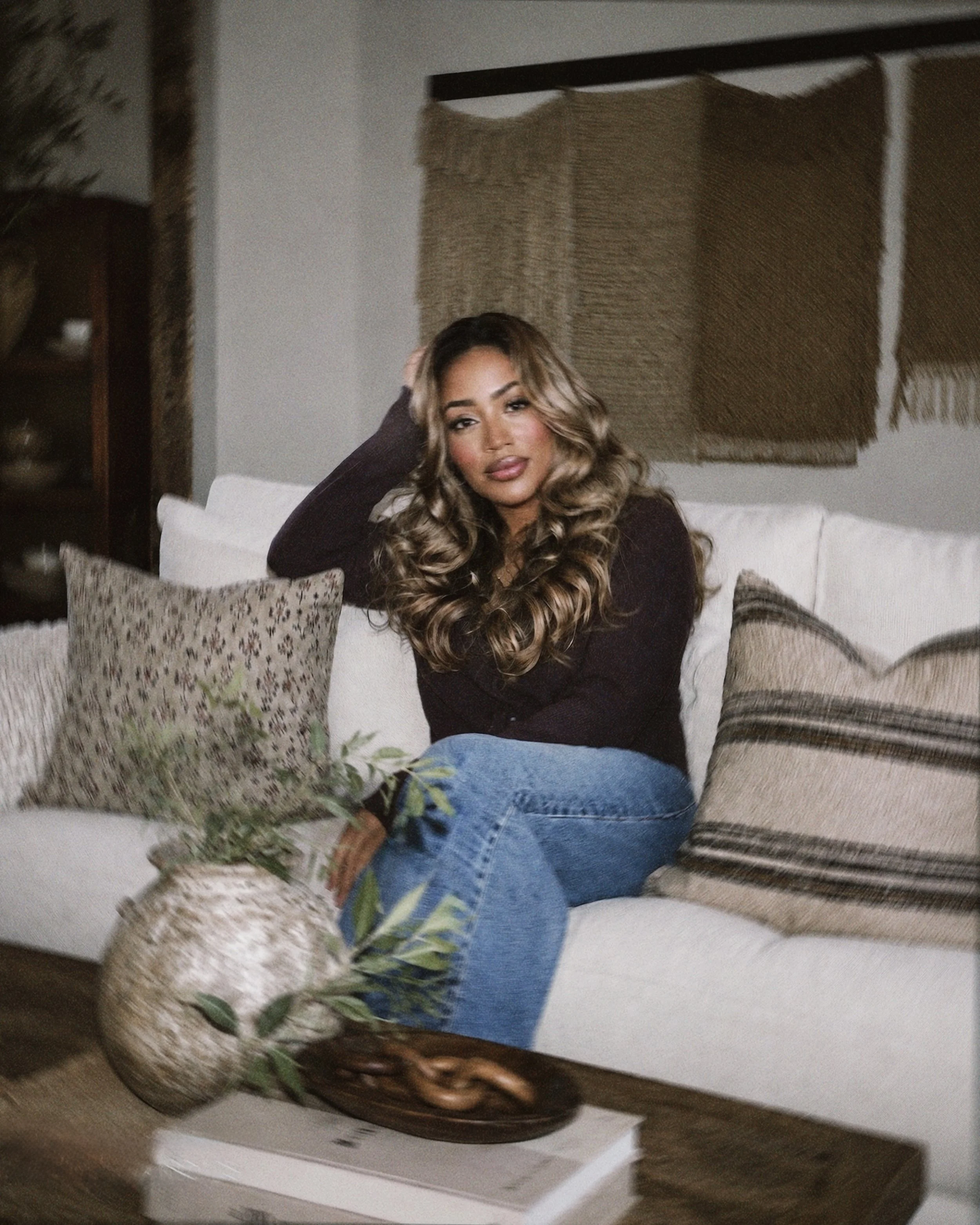 A woman with long, wavy brown hair and wearing a dark long-sleeved top sits on a couch with her left arm resting on her head, looking at the camera. The scene appears to be in a living room with decorative pillows, a ceramic vase, and a mirrored wall hanging in the background.