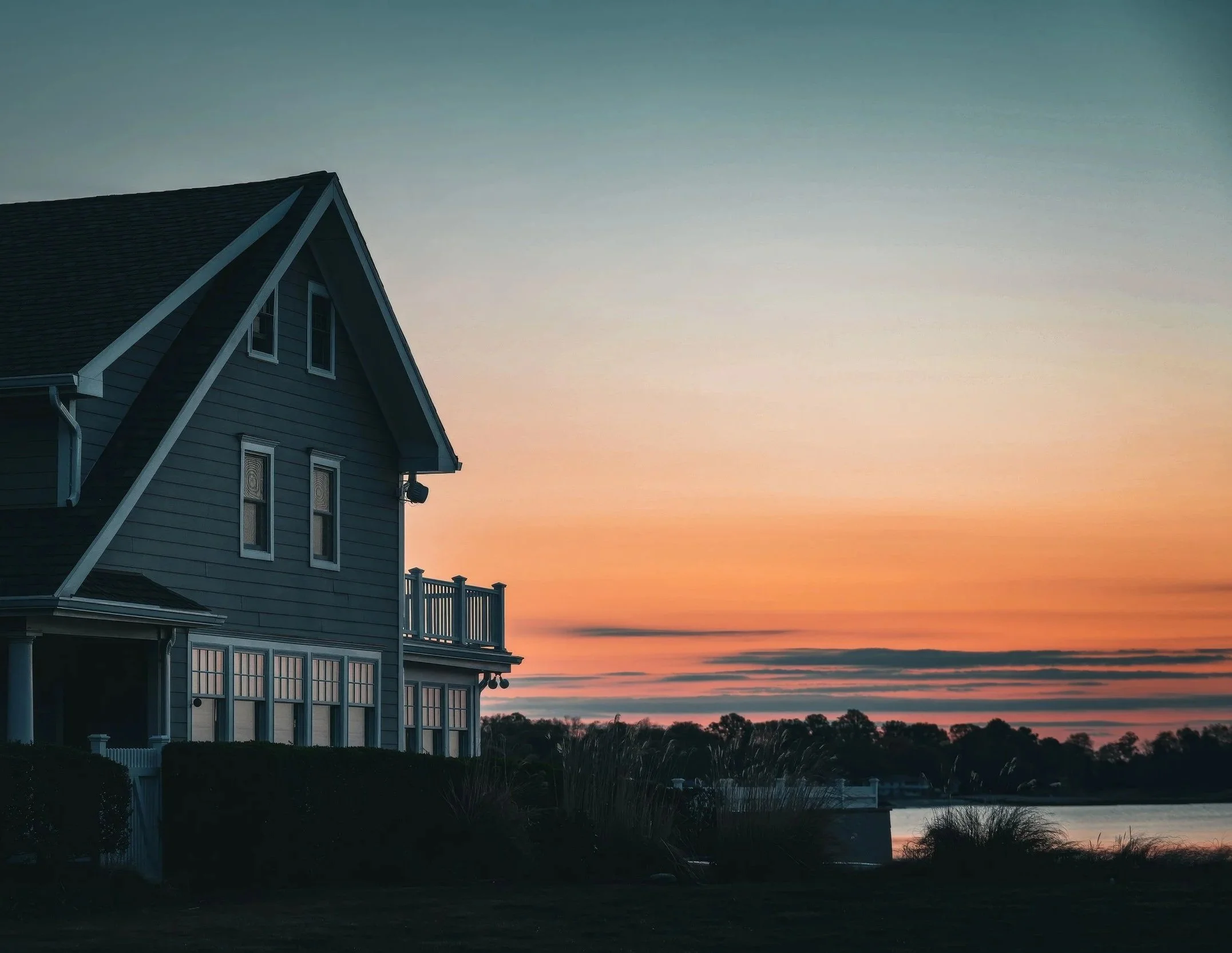 A large, two-story house with blue siding and a steep gabled roof seen at sunset, overlooking a body of water with trees in the distance.