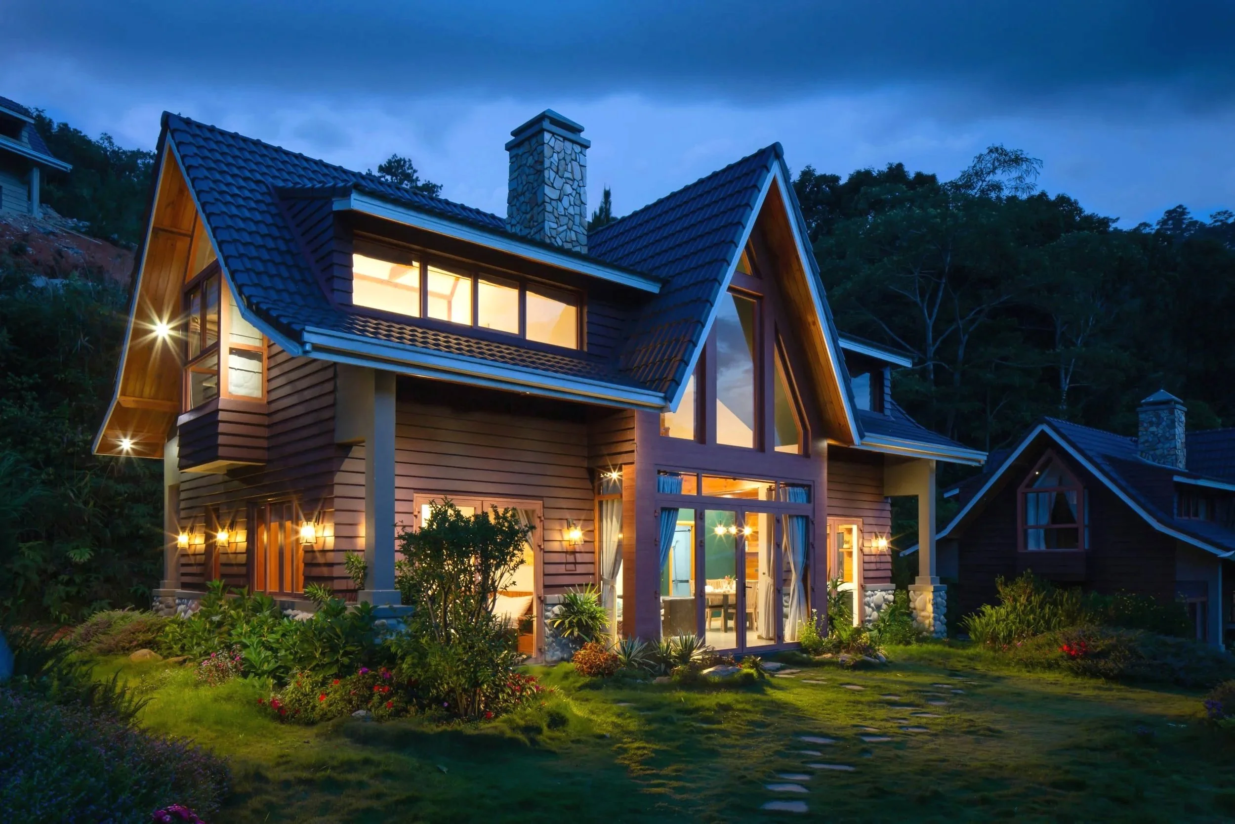 A modern house with large glass windows illuminated from inside, surrounded by a well-maintained garden with flowers and shrubs, set against a dark evening sky.