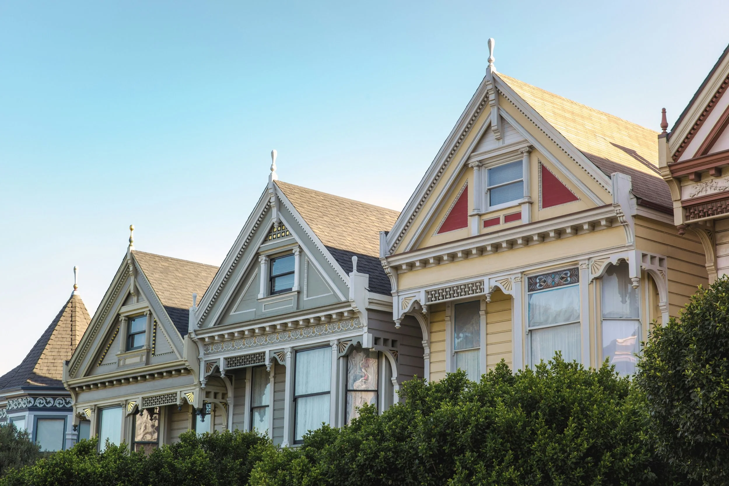 A row of Victorian-style houses with ornate trimming, gabled roofs, and large windows, surrounded by green bushes under a clear blue sky.