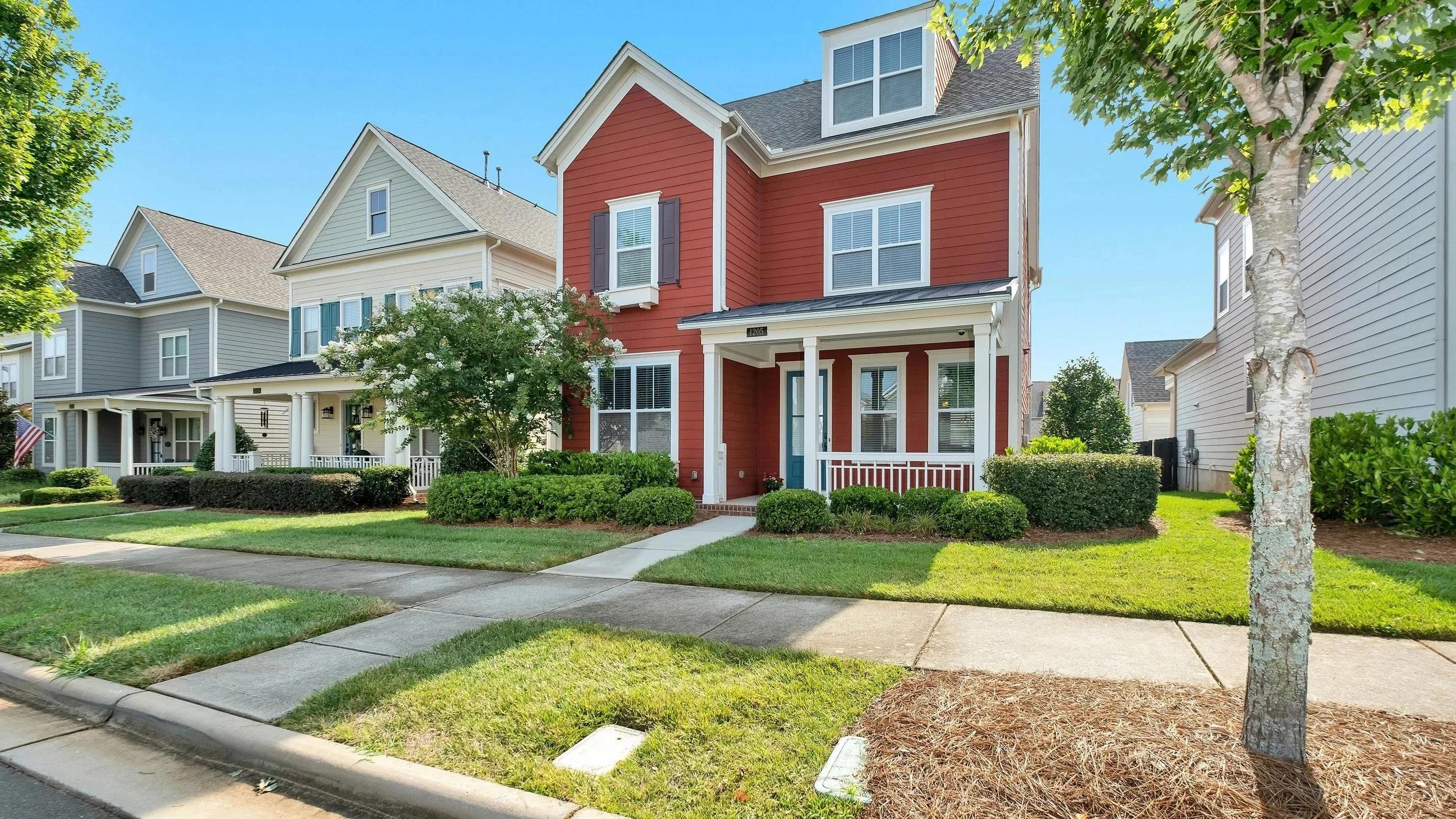 A red two-story house with white trim and a small front porch, surrounded by green bushes and trees, in a neighborhood with similar houses under a clear blue sky.