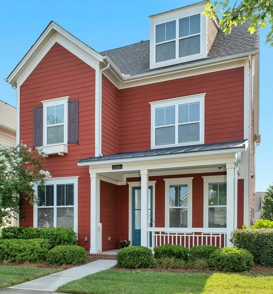 Front view of a red, two-story house with white trim, purple shutters, a small porch with a white railing, a blue front door, and landscaped bushes and lawn.