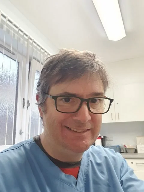 Martin Giebner, Advanced Nurse Practitioner, with glasses, smiling in a clinic room, wearing blue medical scrubs.