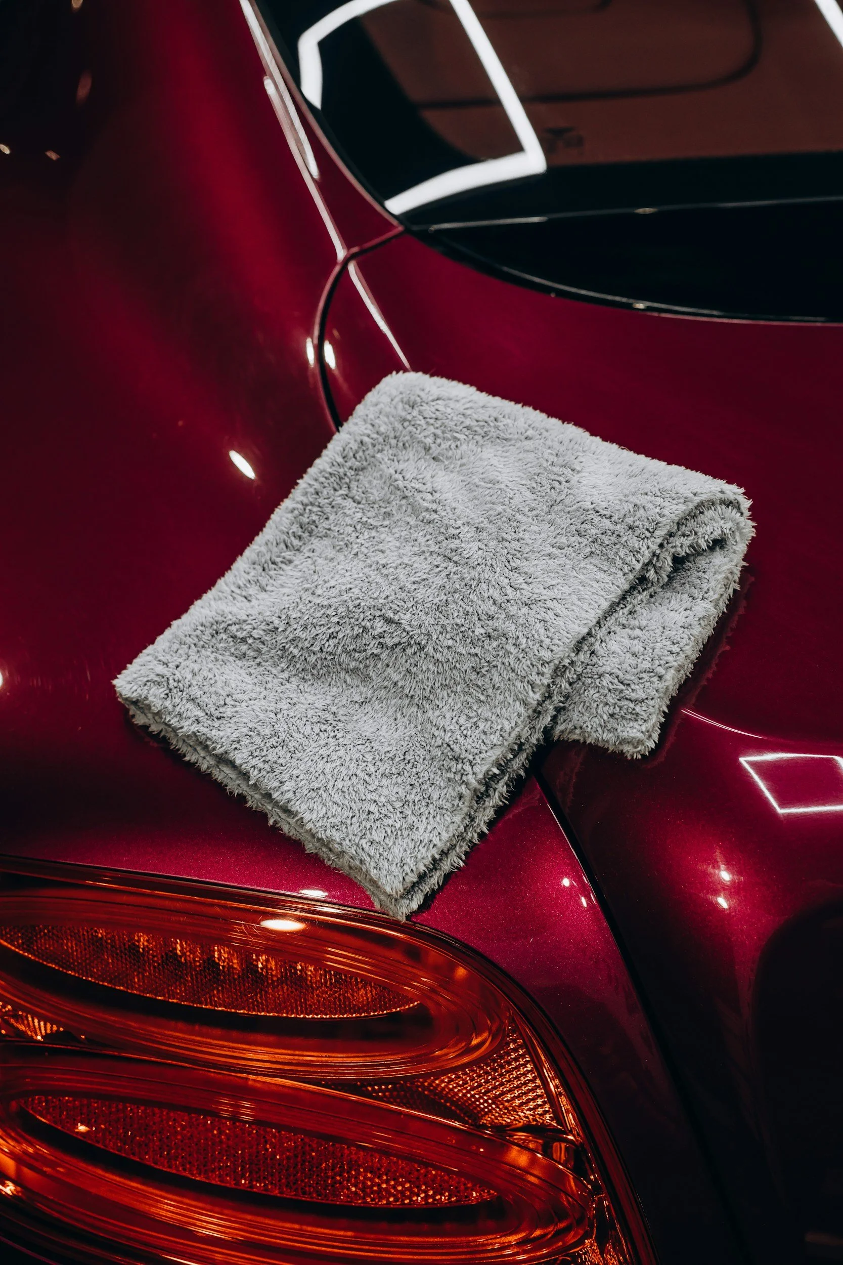 Close-up of a red car's rear with a folded gray towel placed on the trunk, showing reflections and black detailing around the window.