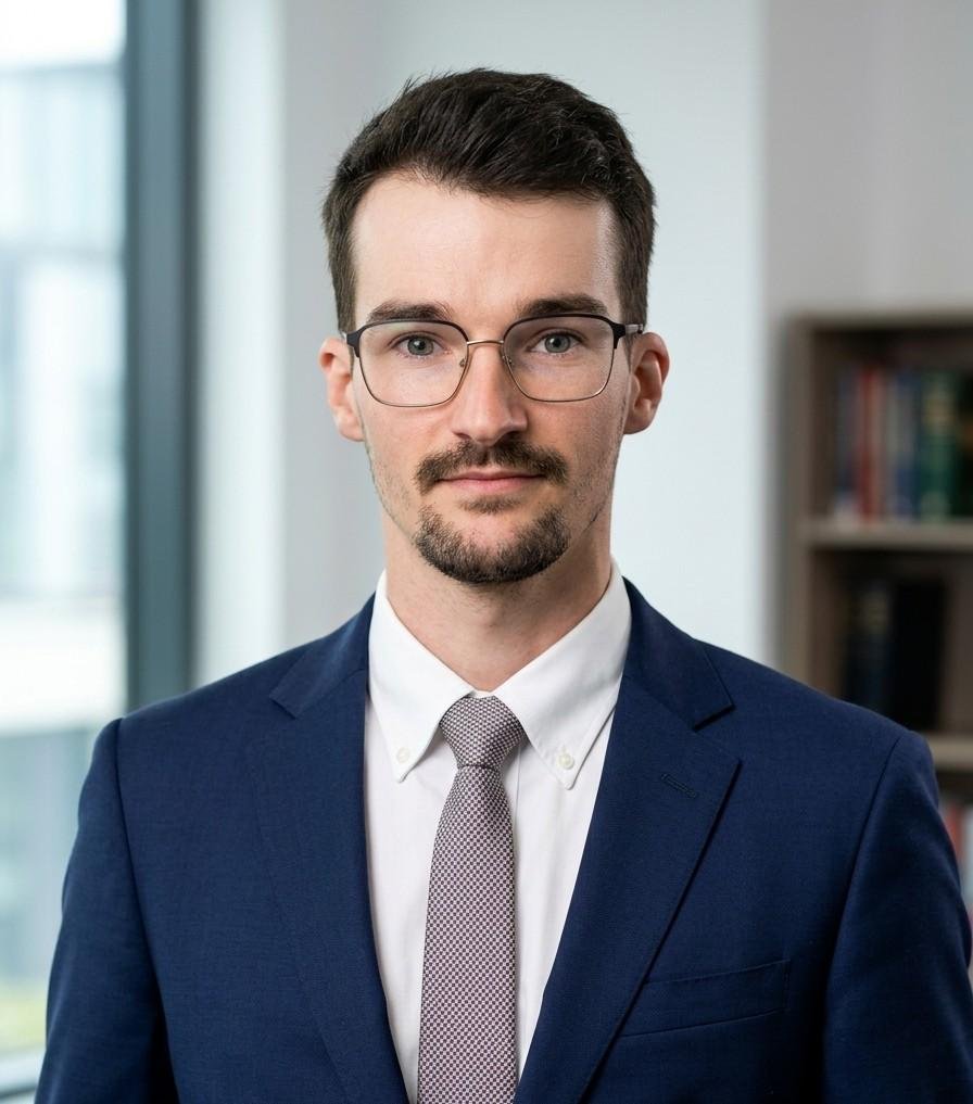 A man with short dark hair, glasses, and a beard, wearing a navy suit, white shirt, and light gray tie, standing indoors near a bookshelf and window.