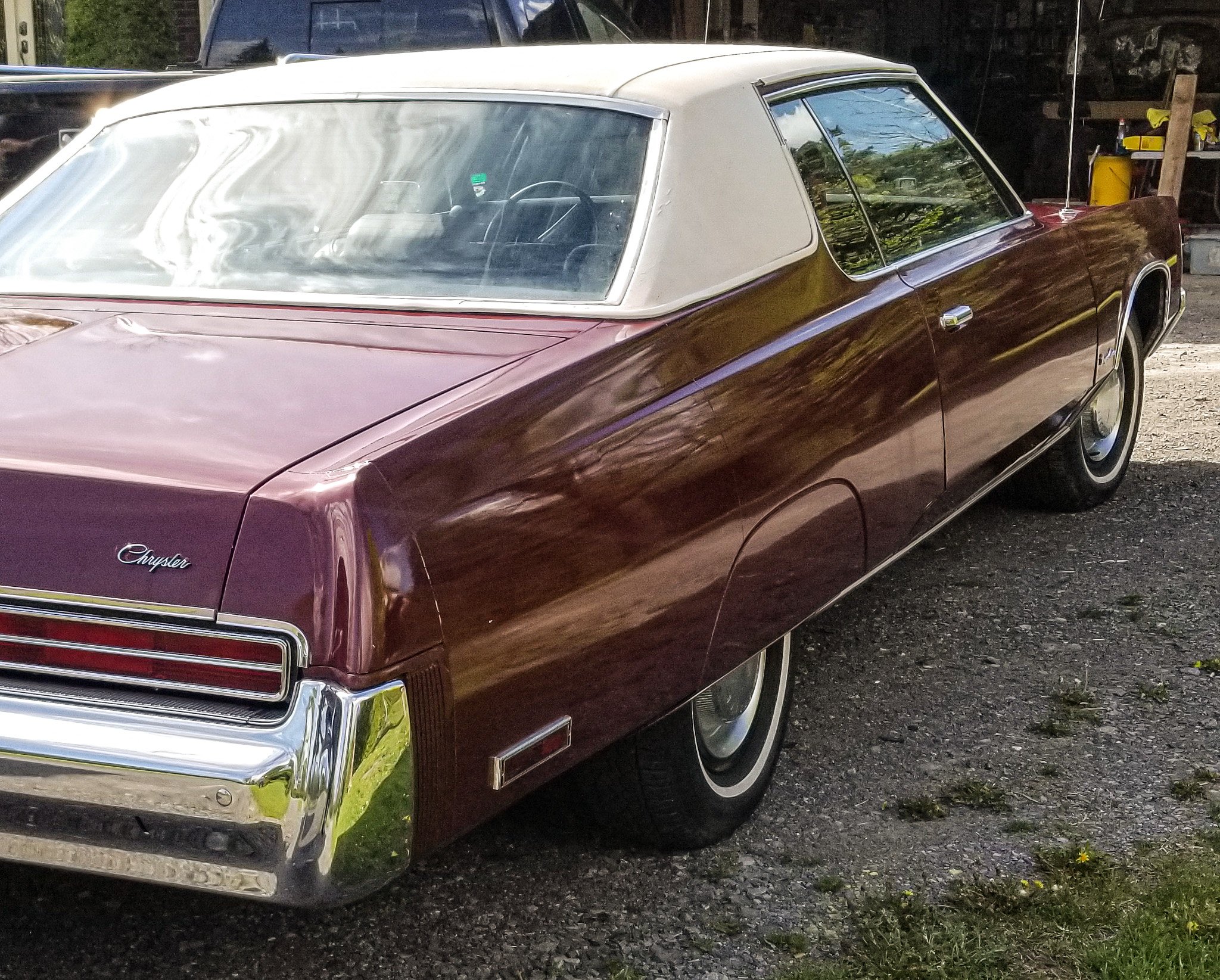 A vintage Chrysler car with a maroon body and white roof parked outdoors on a gravel surface. The car has chrome accents and a distinctive front grille, with trees and a shed visible in the background.