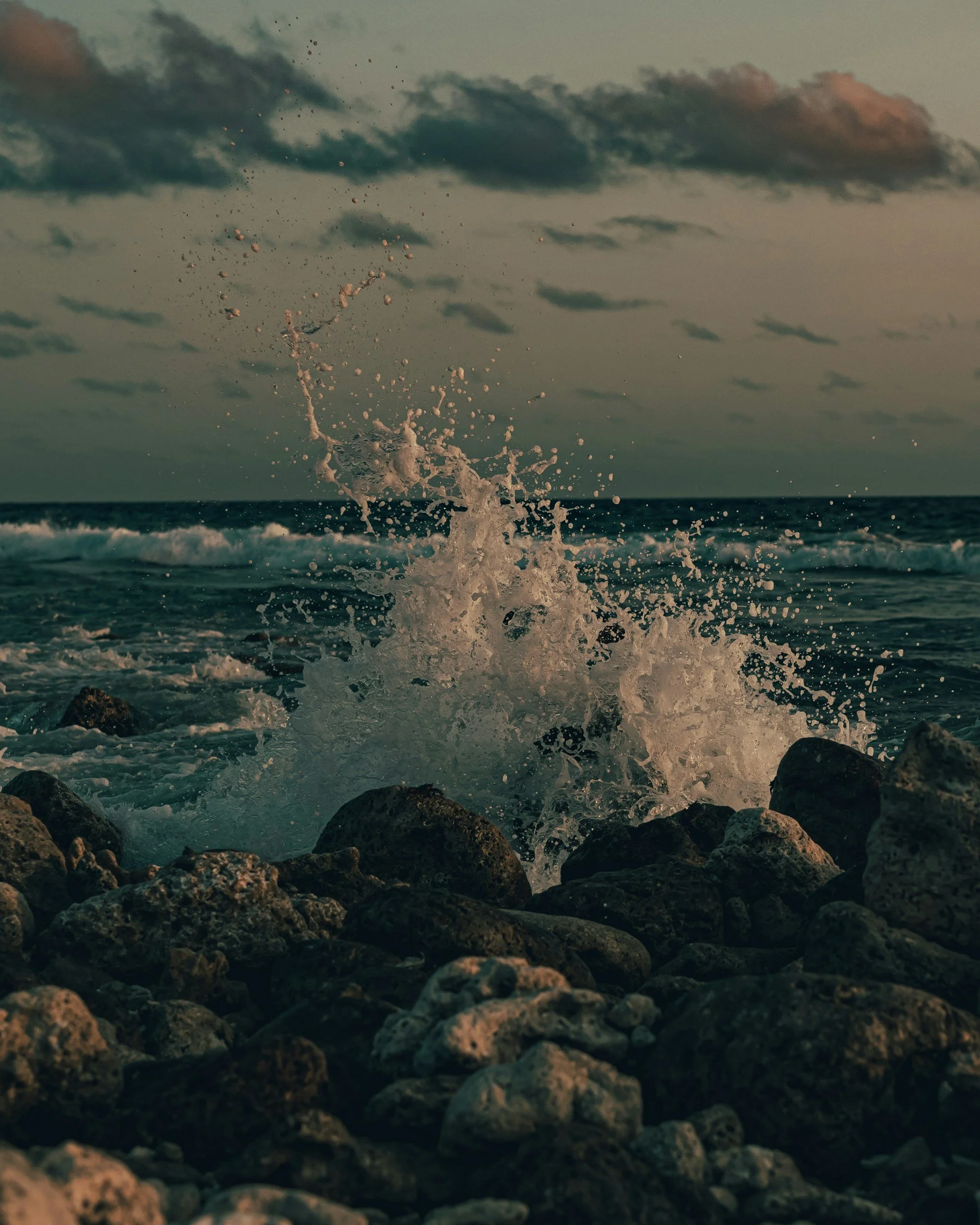 Waves crashing against rocks on a beach with dark clouds in the sky during sunset or dusk.