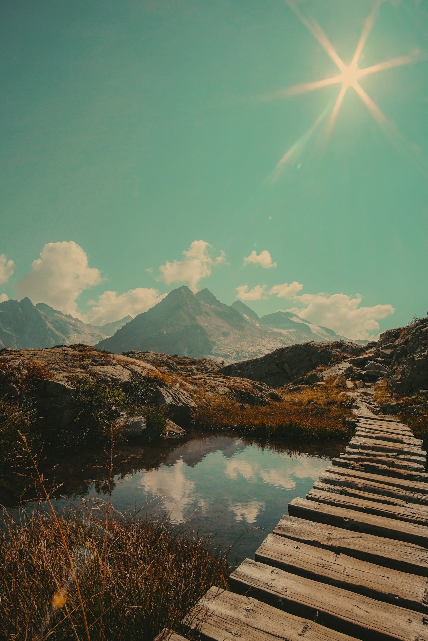 Mountain landscape with a wooden boardwalk, a reflective stream, rocky terrain, mountains in the background, partly cloudy sky, and a bright sun.