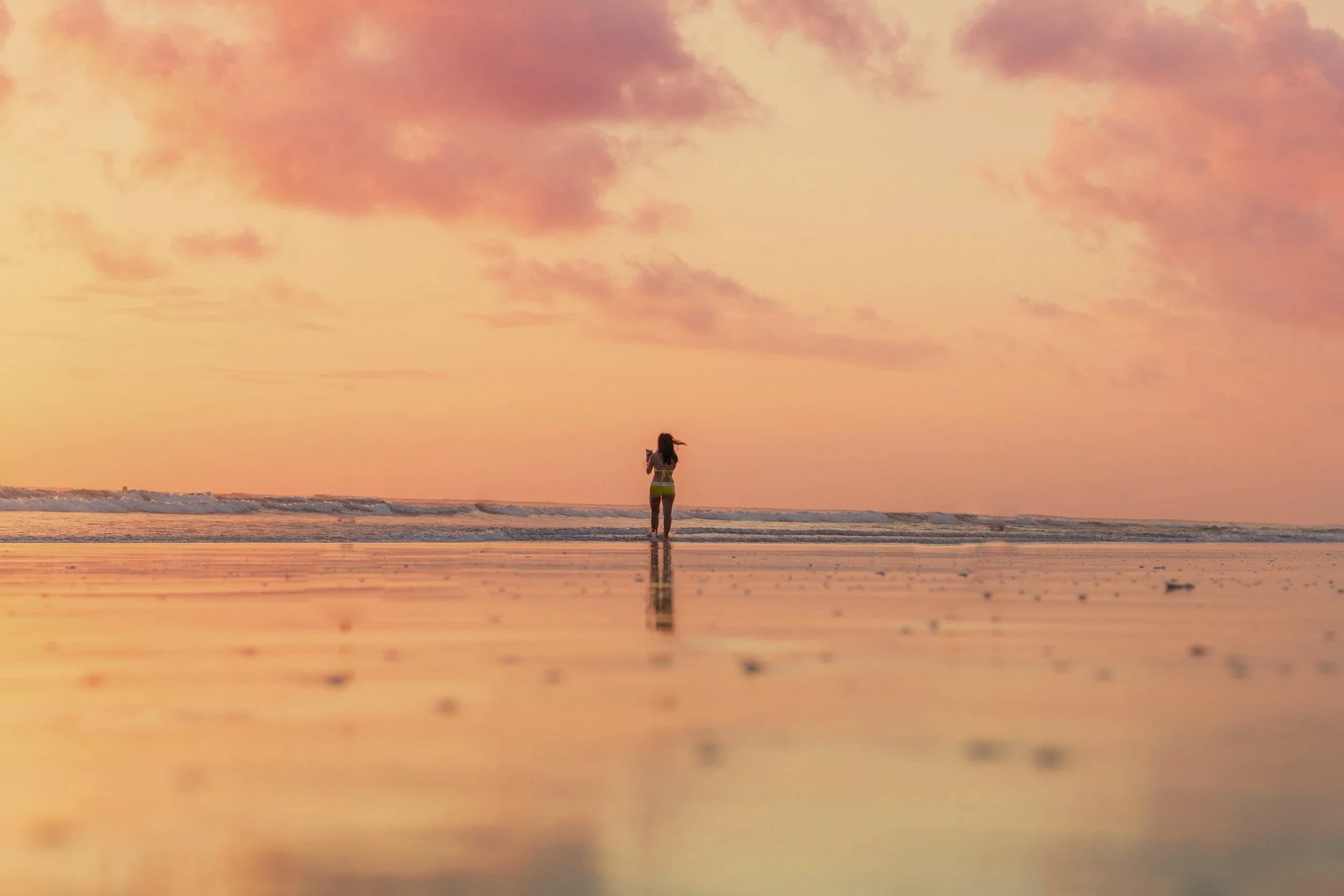 A person stands at the shoreline during a sunset or sunrise, with the sky painted in shades of pink, orange, and purple, reflecting on the wet sand.