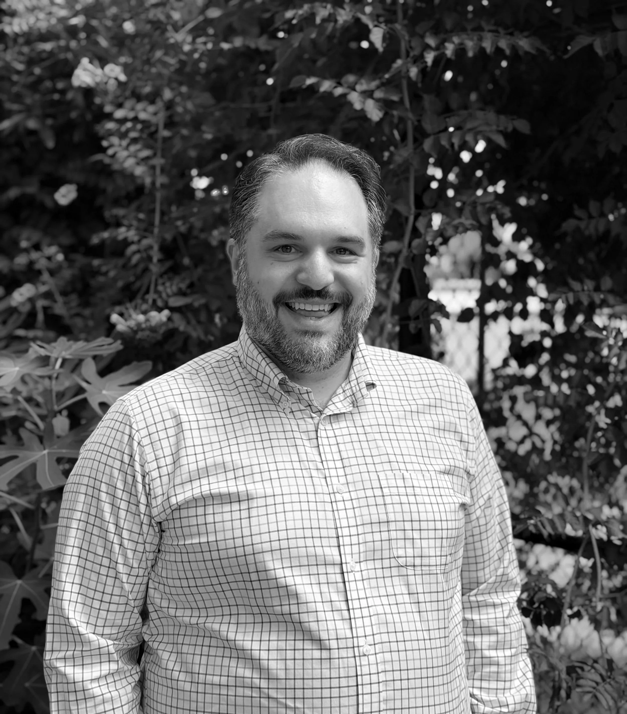 Dr. Mark Versella, a clinical health psychologist who specializes in therapy for healthcare professionals and people with medical and mental health challenges. He has a beard and wears a checked shirt outdoors in front of leafy bushes.