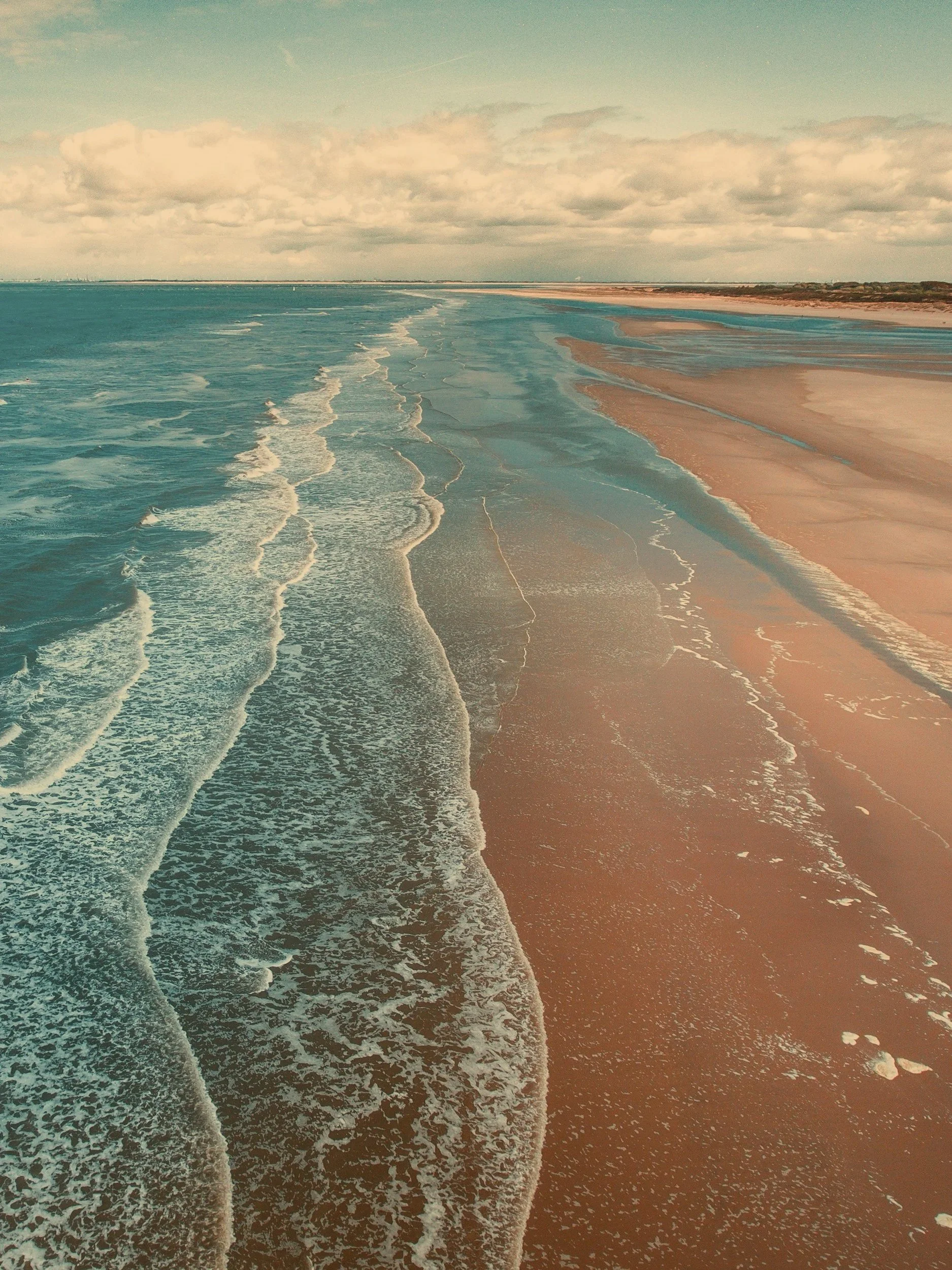 A scenic view of a beach with waves crashing onto the sandy shore under a partly cloudy sky.