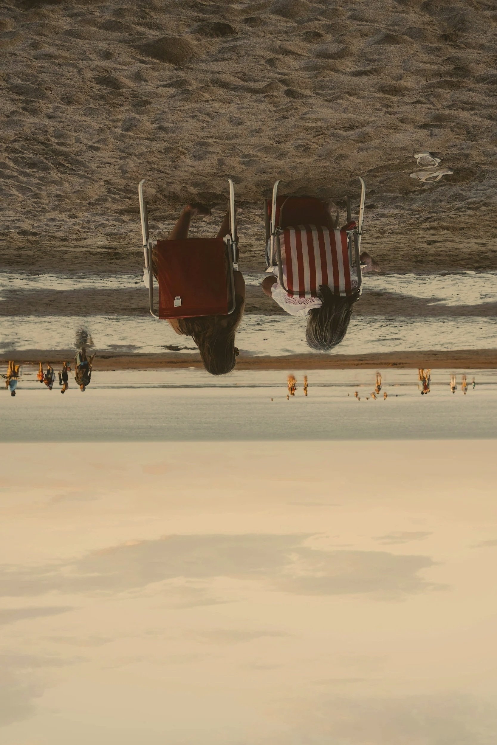 Two people sitting on beach chairs, facing the ocean at sunset, with others in the background walking along the shoreline.