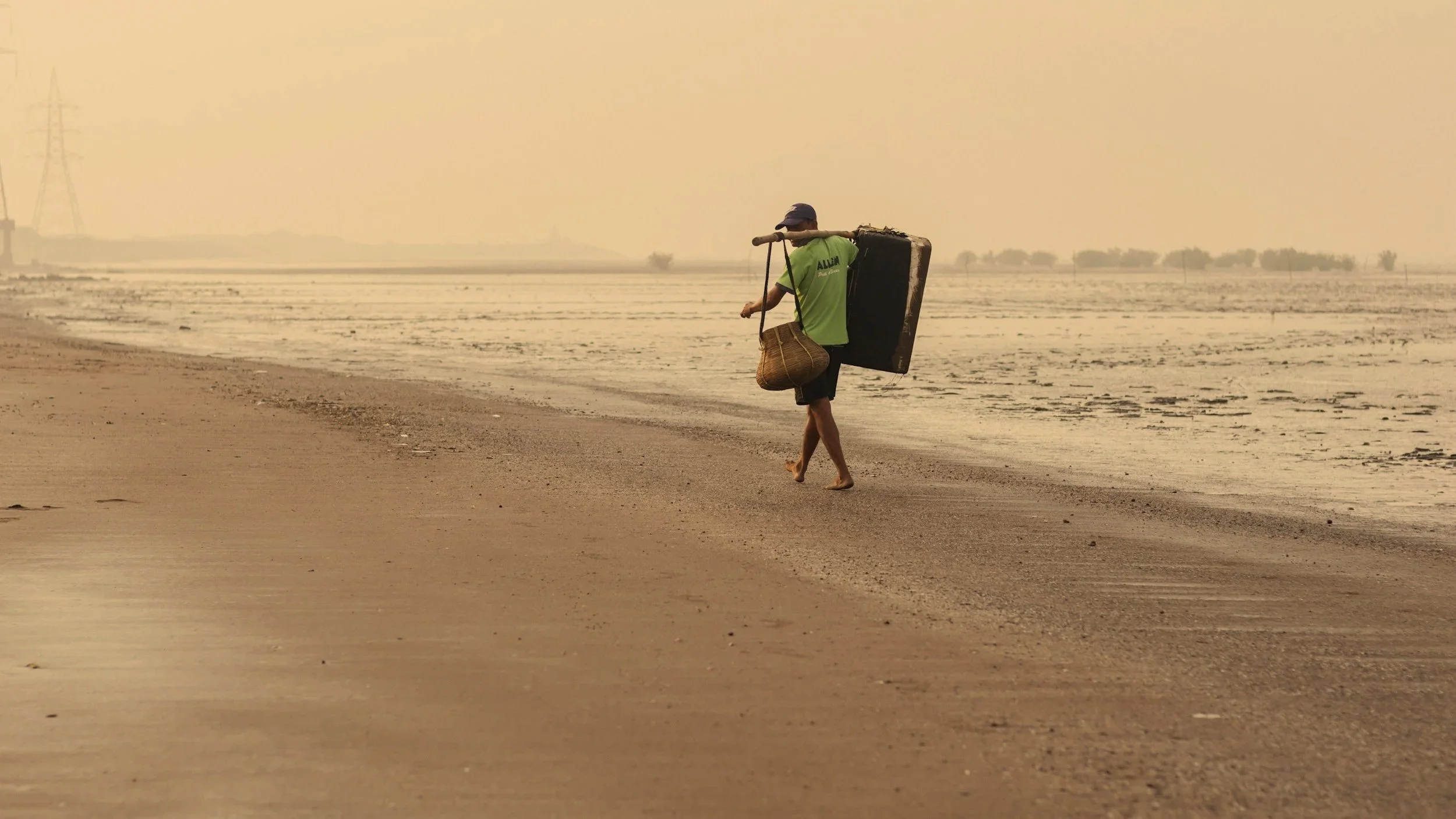 A person walking barefoot along a sandy beach, carrying a large black object on their back with a pole, and holding a basket, with a hazy sky and sparse trees in the background.