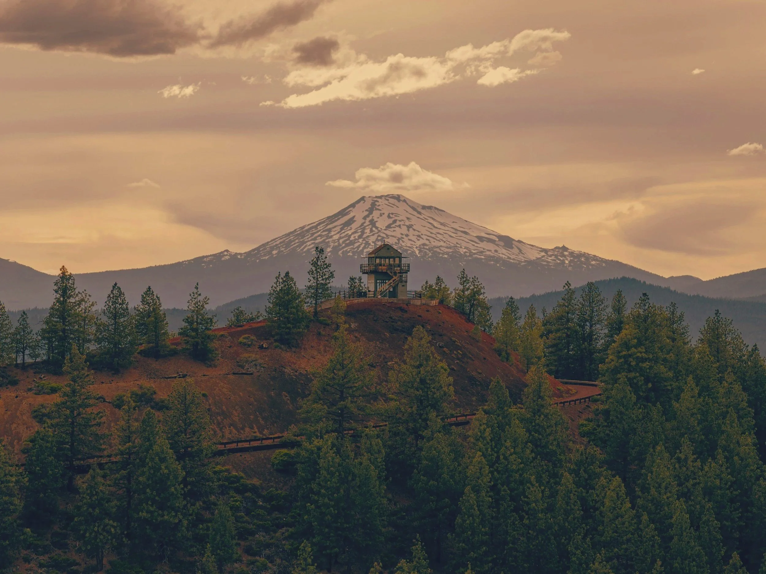 A snow-capped mountain with a small observation building on a hill in front, surrounded by pine trees and a cloudy sky.