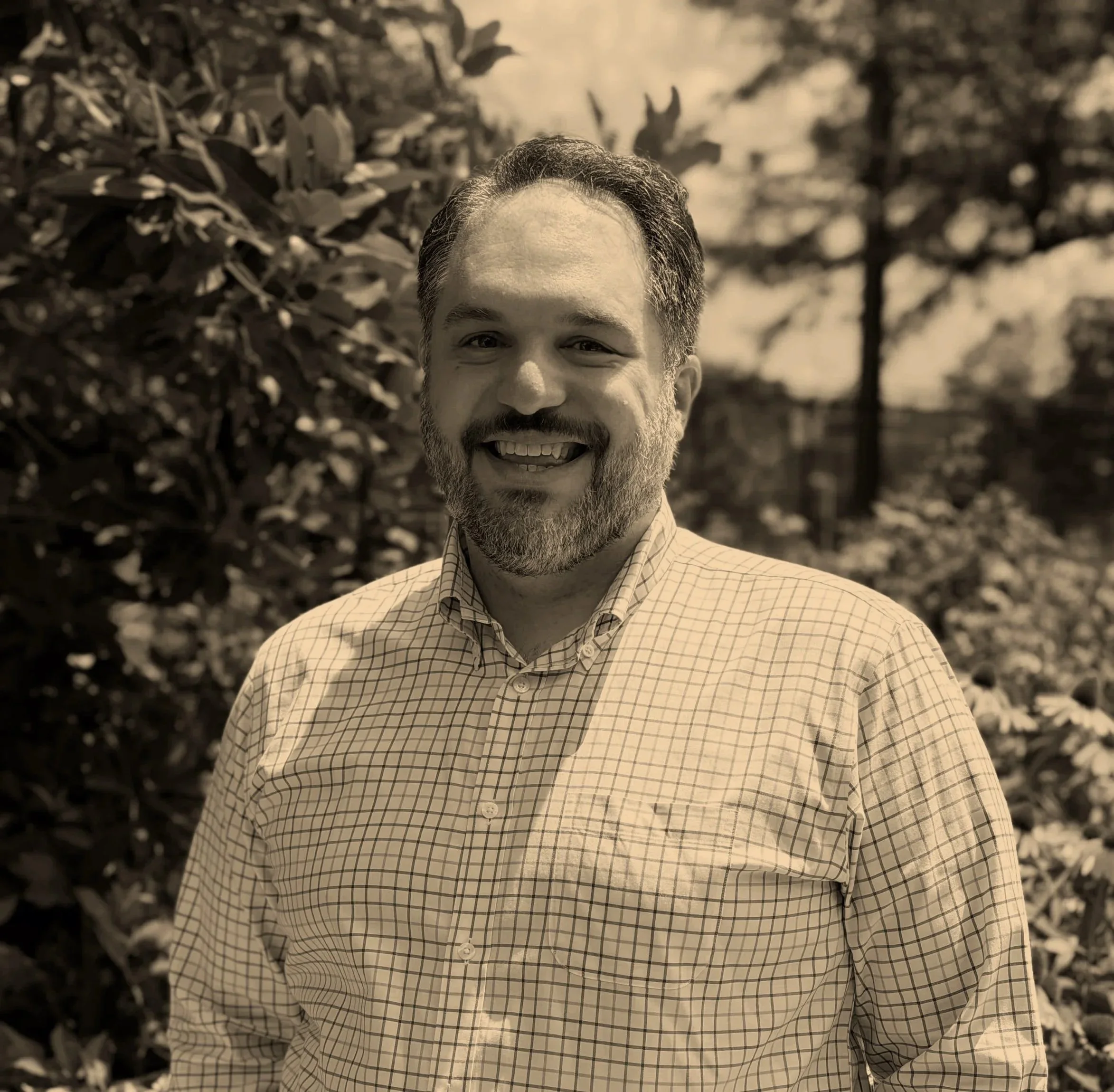 Dr. Mark Versella, a clinical health psychologist who specializes in therapy for healthcare professionals and people with medical and mental health challenges. He's smiling, has a beard and wears a checked shirt outdoors with trees in the background.