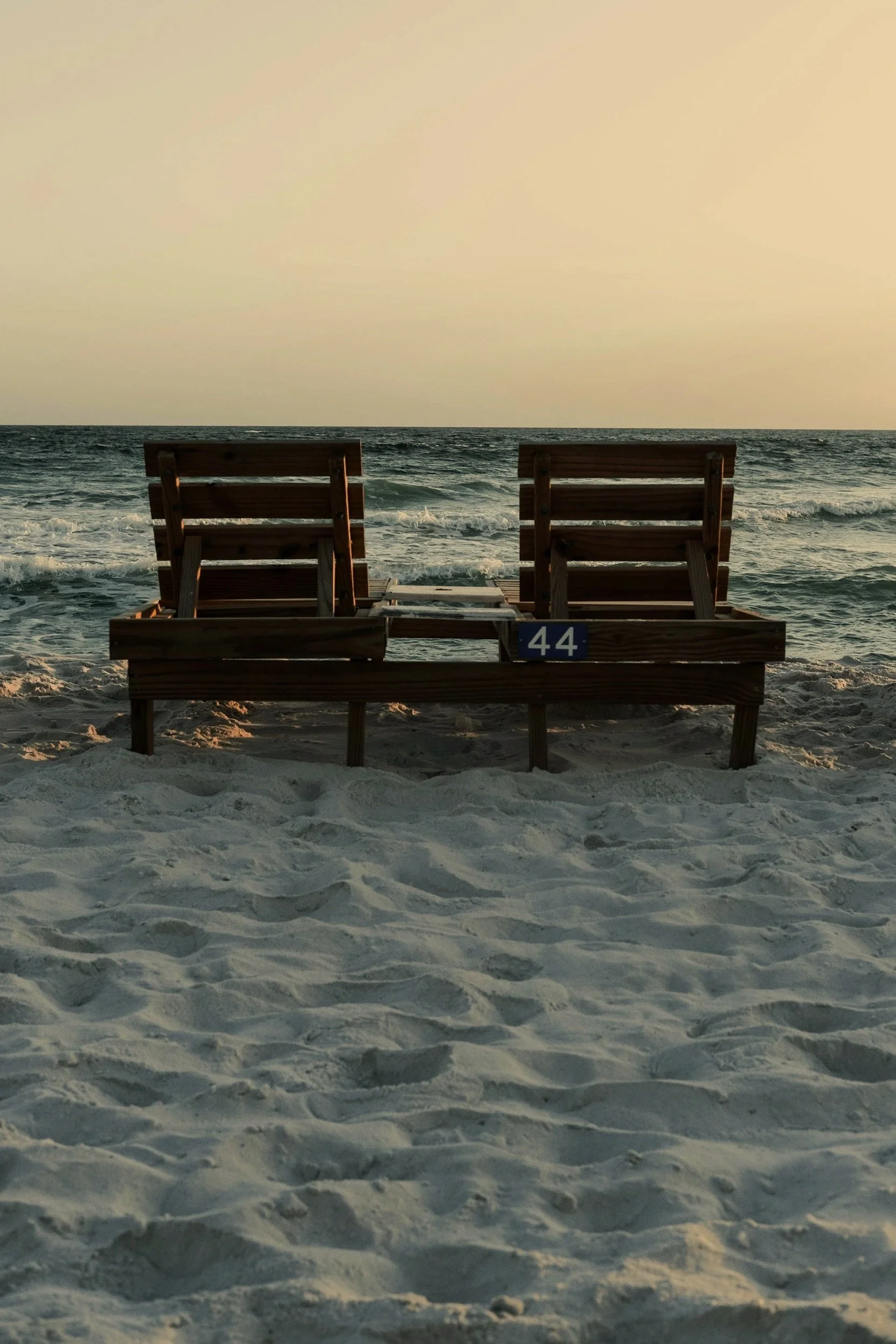 Two wooden lounge chairs on a sandy beach facing the ocean with waves and a cloudy sky.