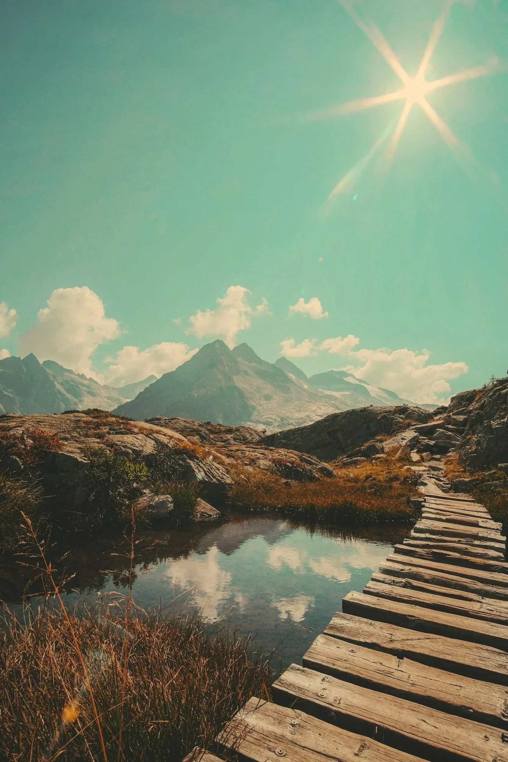A mountain landscape with a clear sky, sun shining, and a small body of water reflecting the mountains. A wooden walkway crosses the foreground.