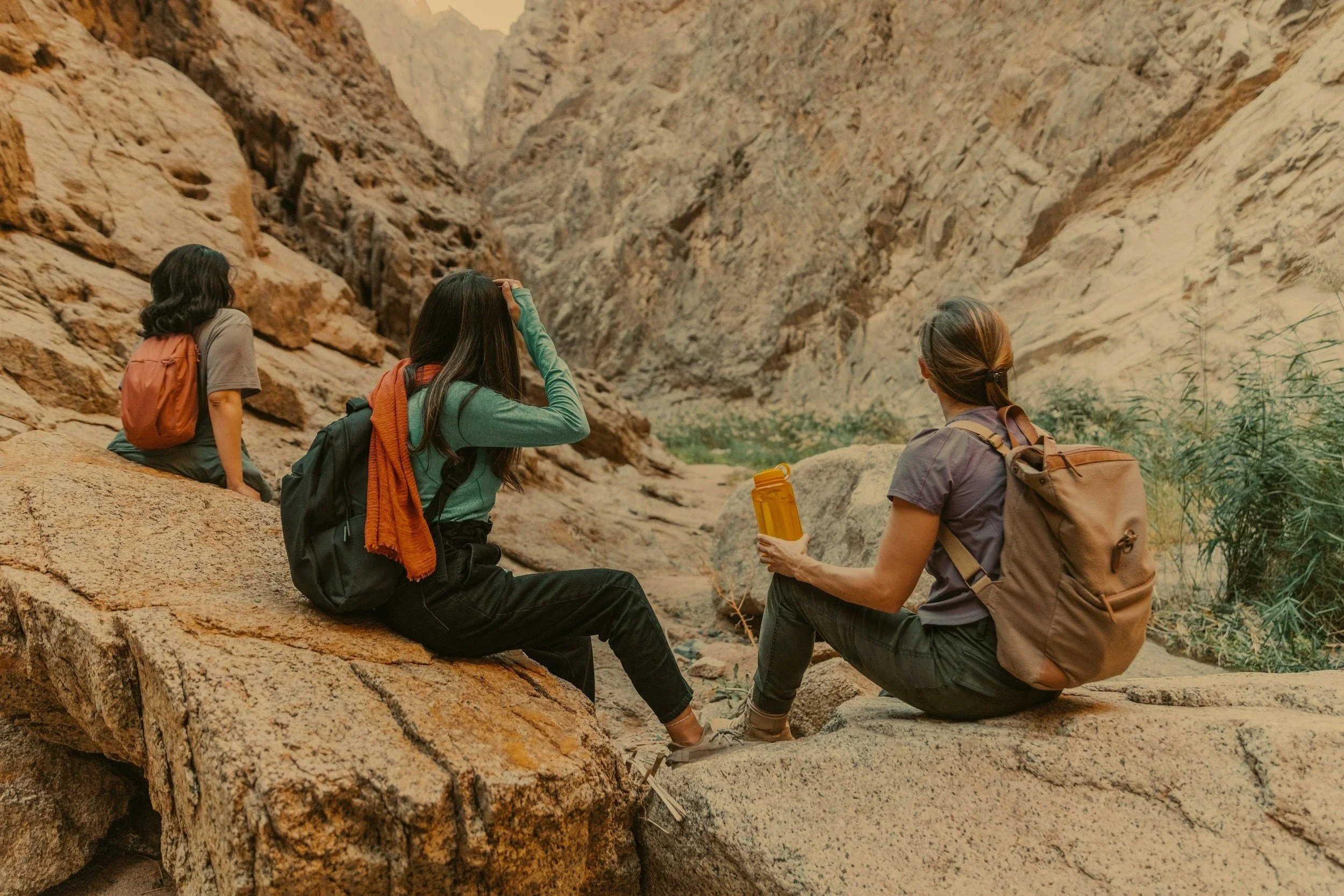Three women with backpacks sitting on large rocks in a canyon, looking at the rocky, arid landscape ahead.