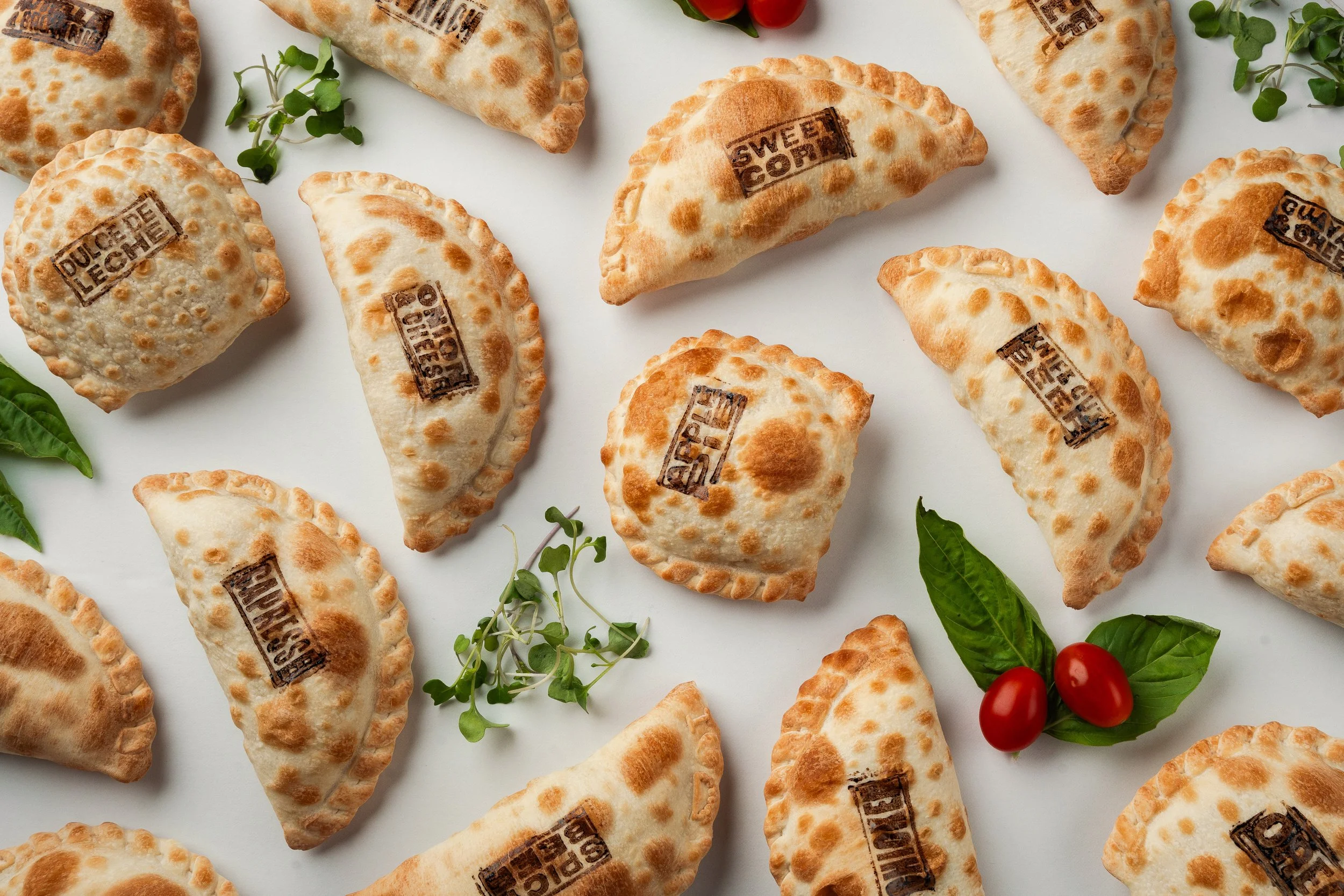 Assorted half-moon shaped empanadas with stamped labels, green leaves, and cherry tomatoes on a white background.