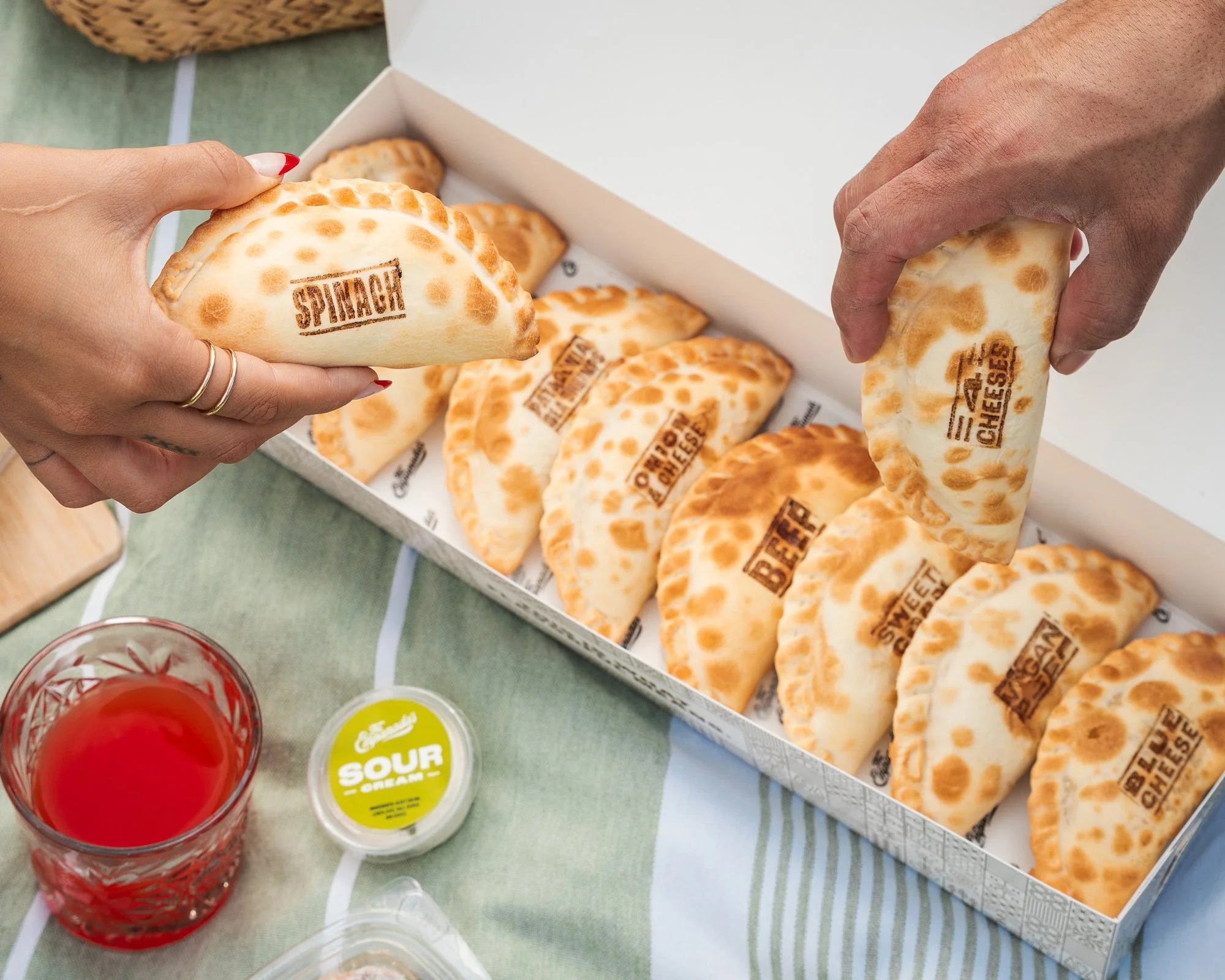 Two hands holding empanadas stamped with the words 'SPOIL ALERT' and 'CHEESE' in a white box, with a glass of red drink and some small containers of condiments on a table.