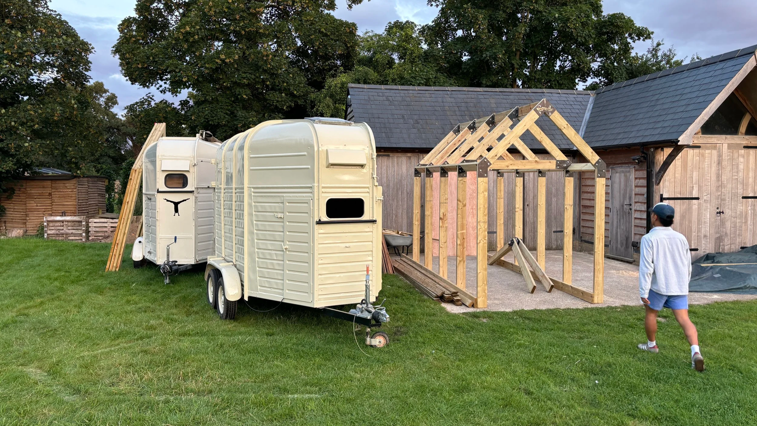 A person standing in a backyard watching a construction site for a wooden shed or extension, with a white trailer and a partially built wooden frame, green grass, trees, and a wooden shed in the background.