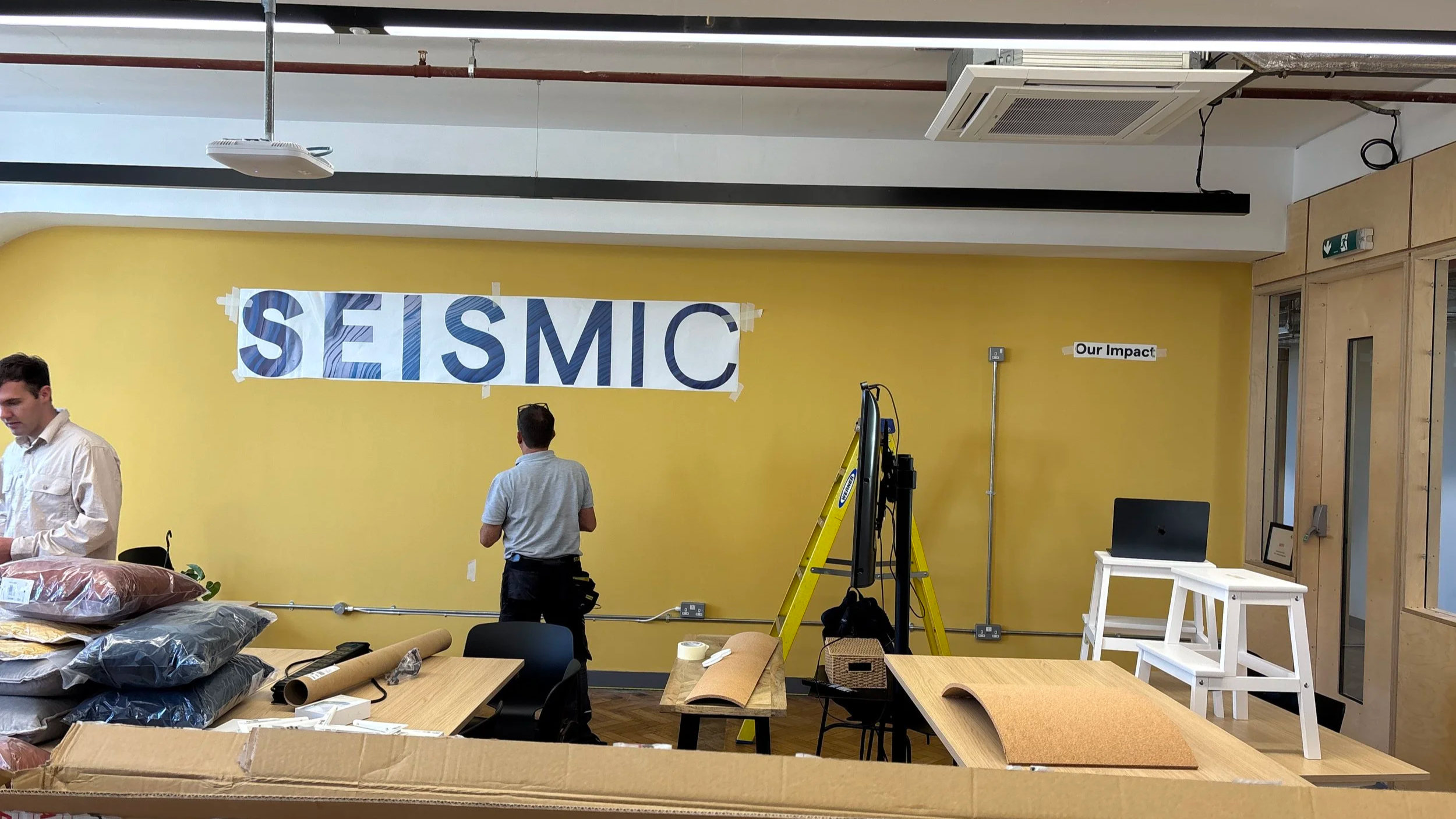 Office space with yellow walls, a large banner reading 'SEISMIC,' a table with supplies, and three people preparing the area, with tools and equipment visible.