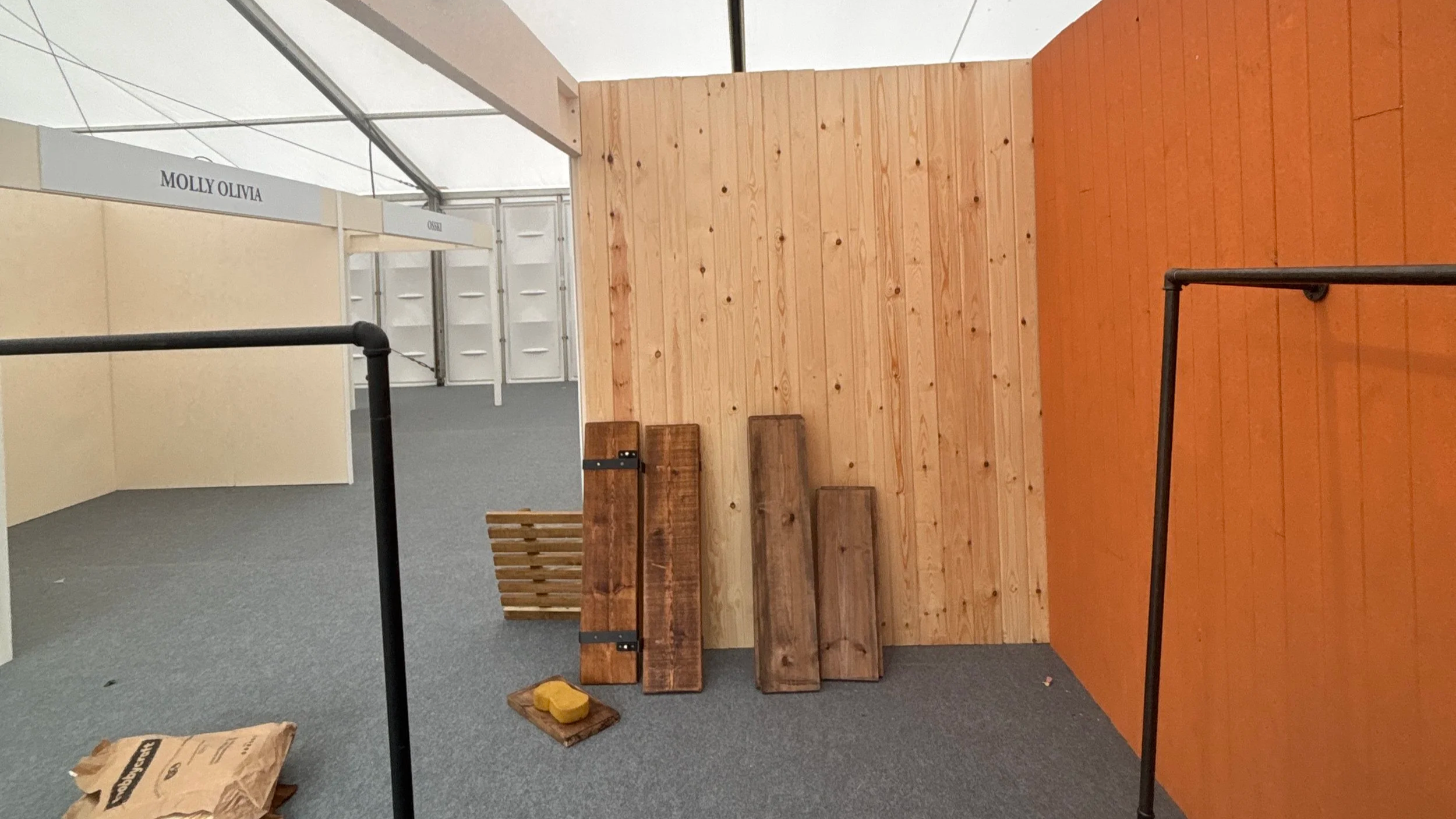 Display of wooden boards and furniture pieces at an indoor exhibition or trade show, with black metal stand in foreground and labeled booths in background.