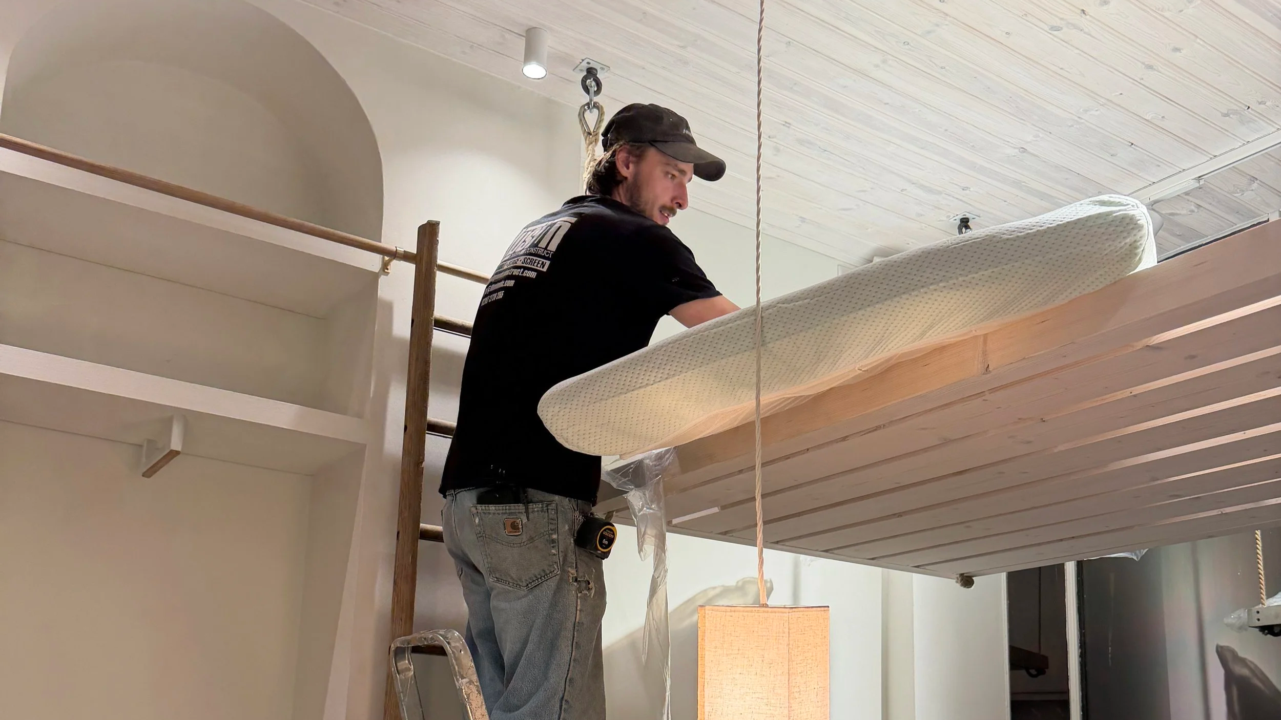 A man wearing a black cap and jeans stands on a ladder, working on a wooden ceiling. He is near a mattress and a climbing rope, in a room with a wooden ceiling and white walls.
