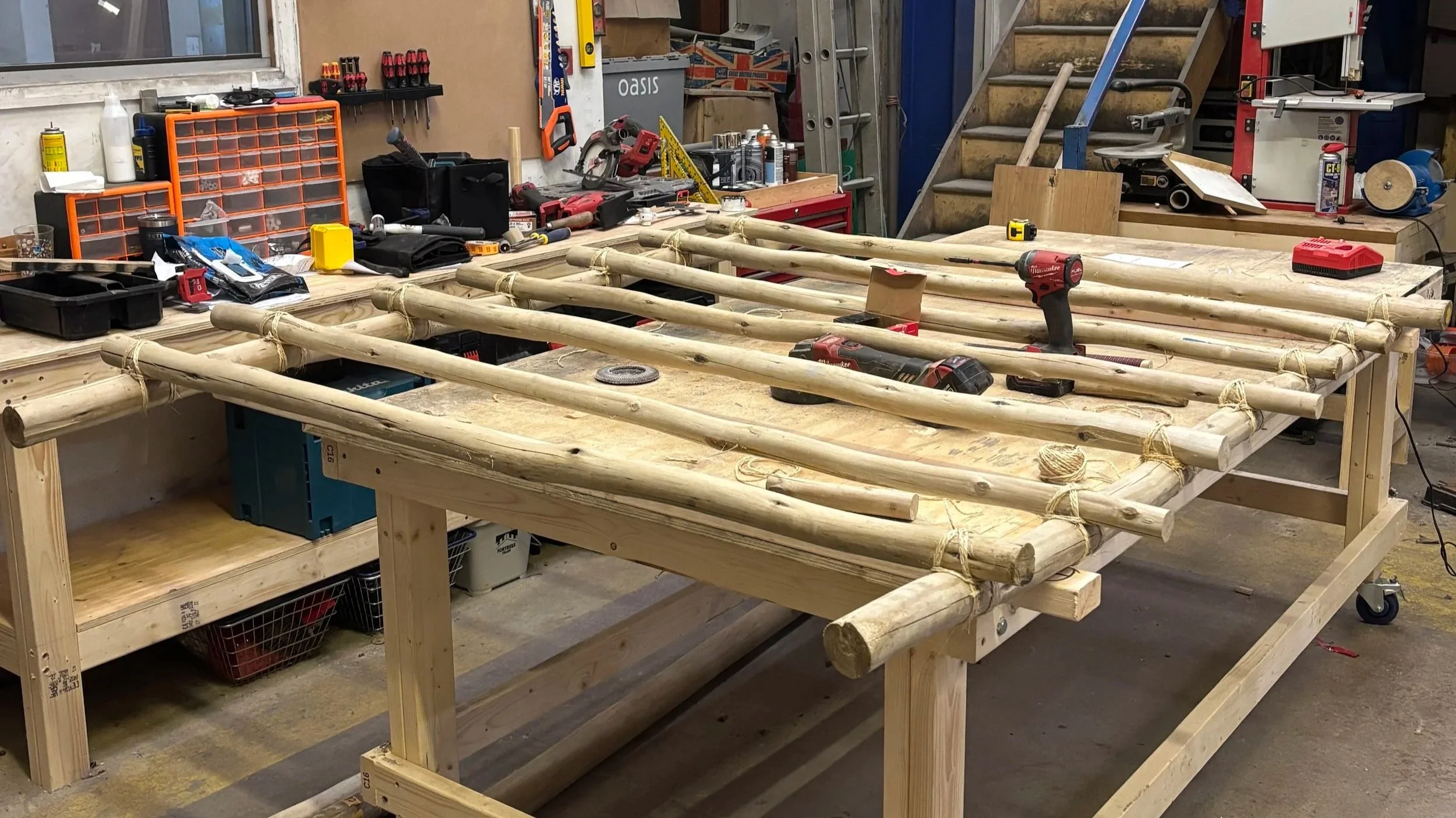 A workshop workspace with a wooden table containing bamboo poles and power tools, surrounded by various tools, supplies, and equipment.