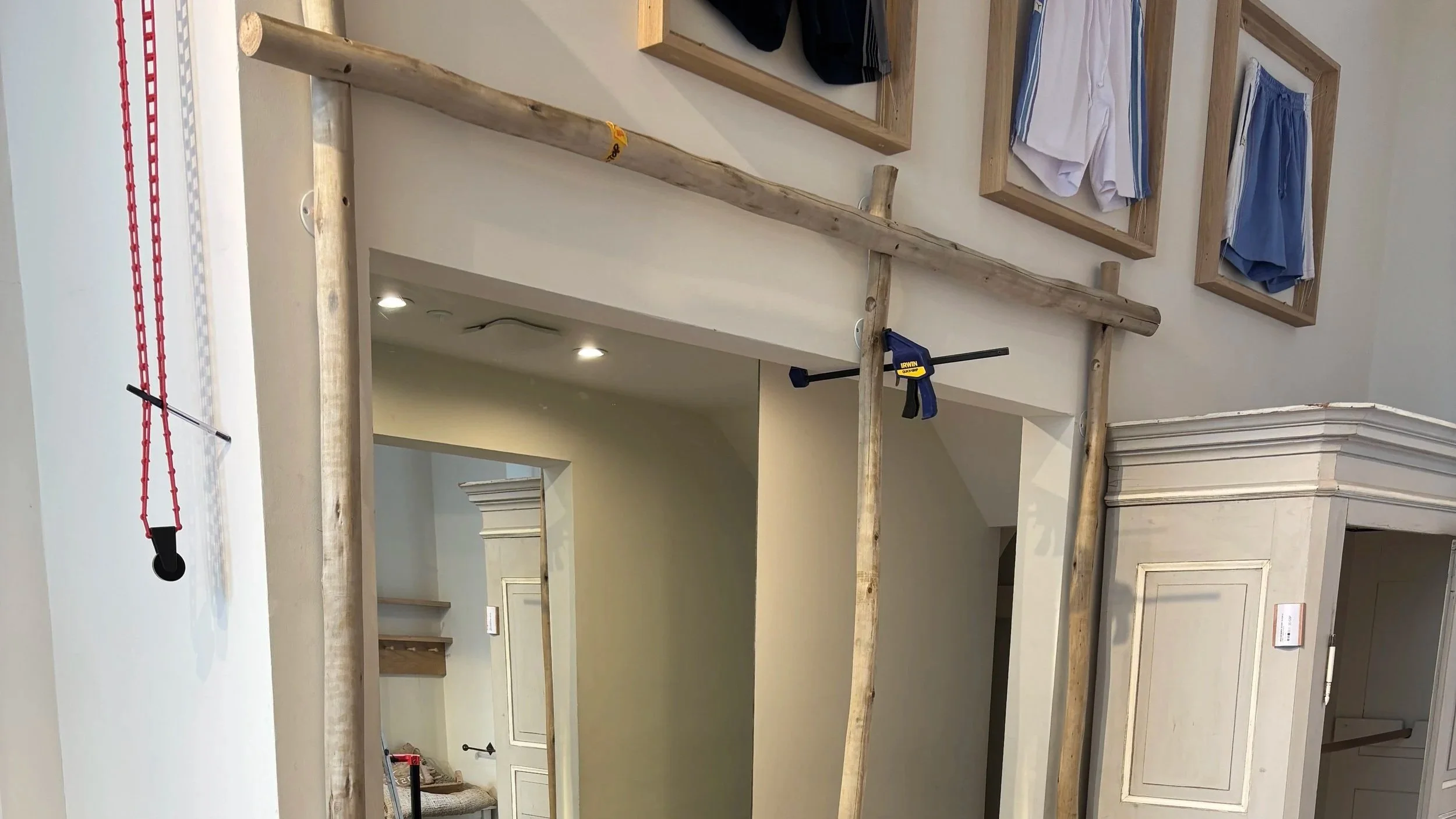 Interior of a house under renovation with wooden beams, a clamp, and a red hanging tool. Clothes are displayed on framed panels on the wall.