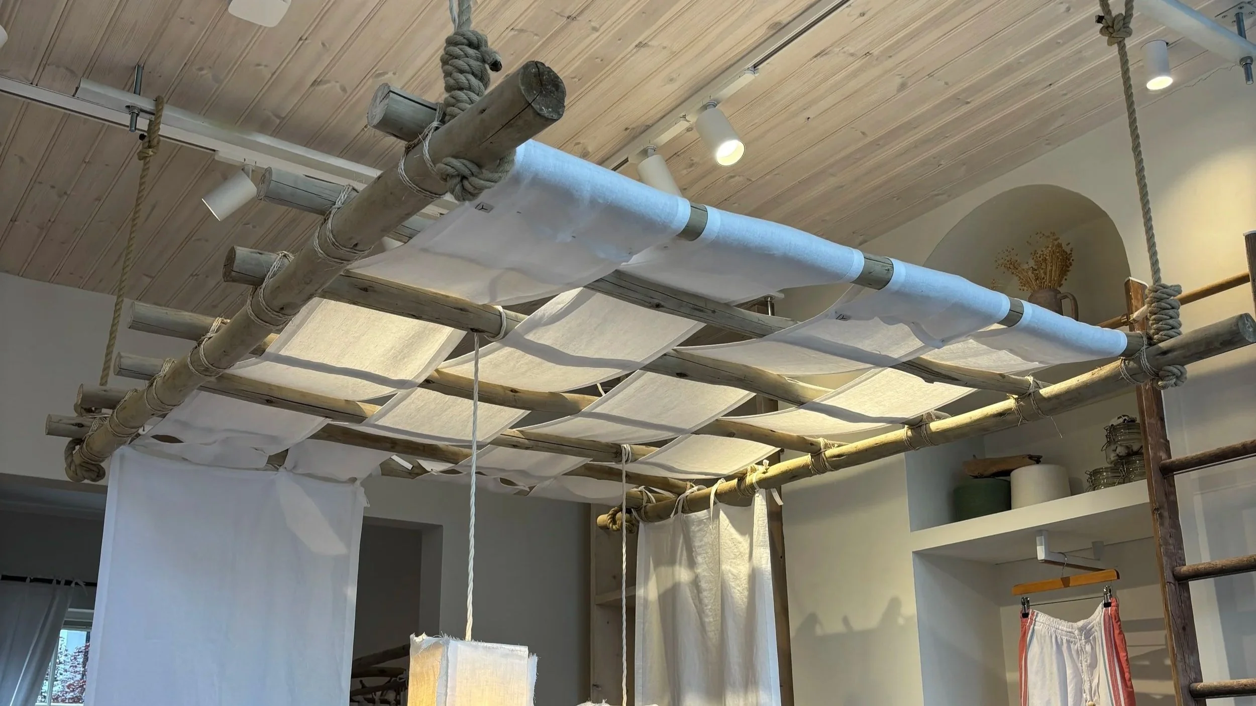View of a cozy indoor space with a ceiling made of wooden planks, featuring a large hanging wooden and rope structure with fabric drapes attached, resembling a makeshift bed or a play area.