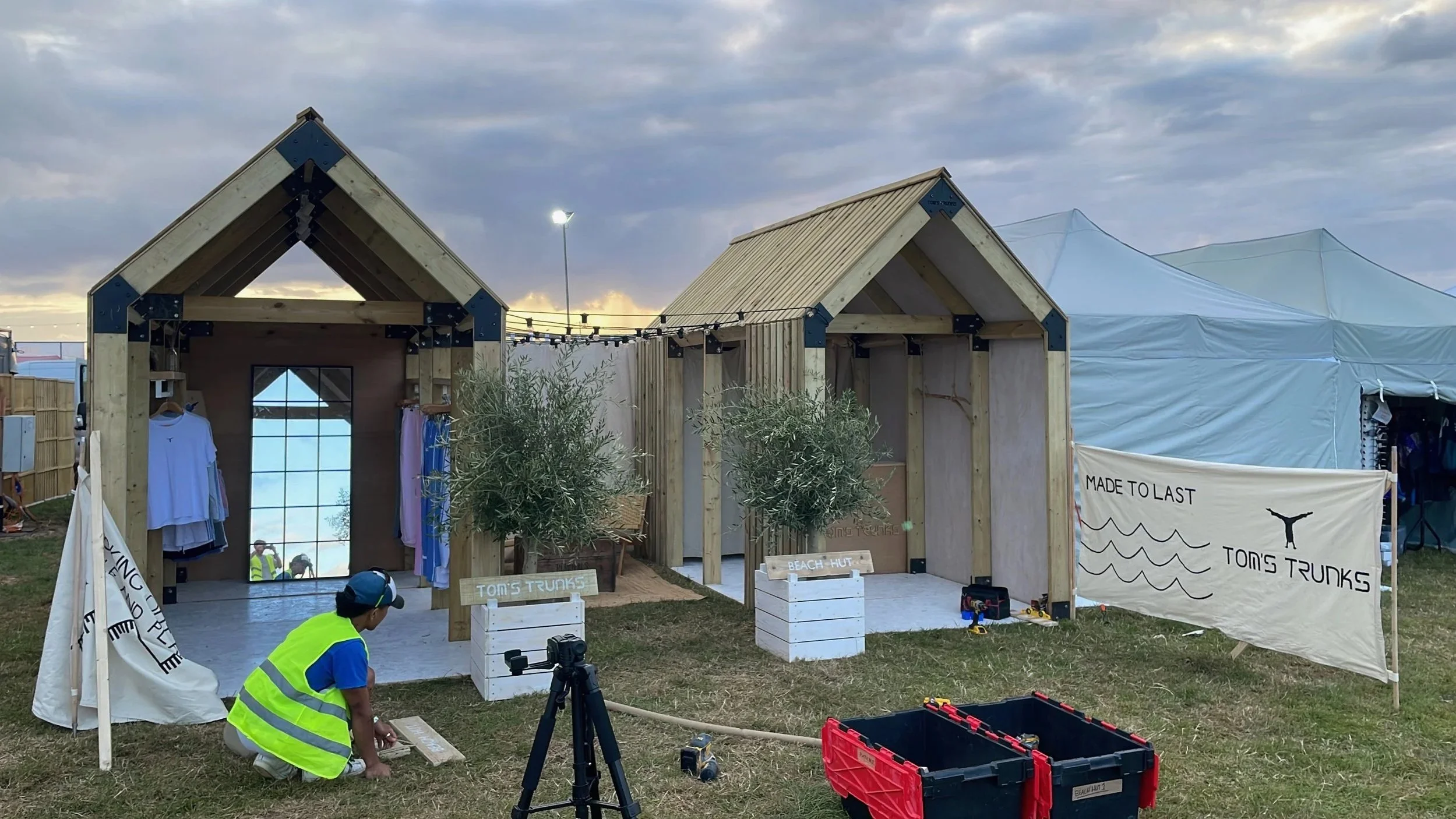 Under construction of a small wooden beach hut with two gabled roofs, white signs reading "Tom's Trunks" and "Beach Hut," surrounded by tools, boxes, and workers in safety vests.