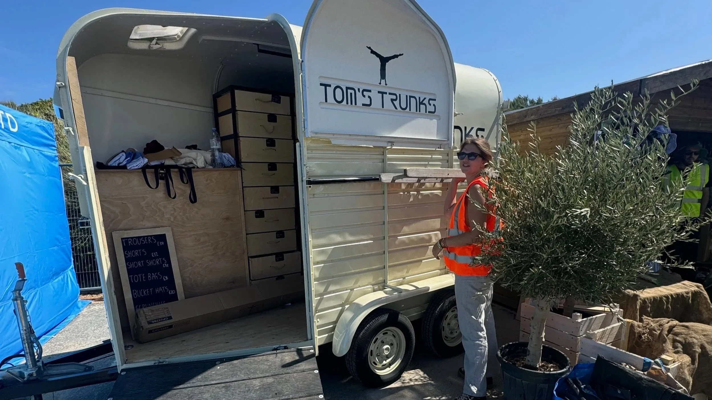 A woman in sunglasses and an orange safety vest stands beside a small white mobile shop named 'Tom's Trunks,' which sells clothing such as trousers and shorts. The shop has an open side revealing shelves with accessories, a chalkboard menu, and a wat