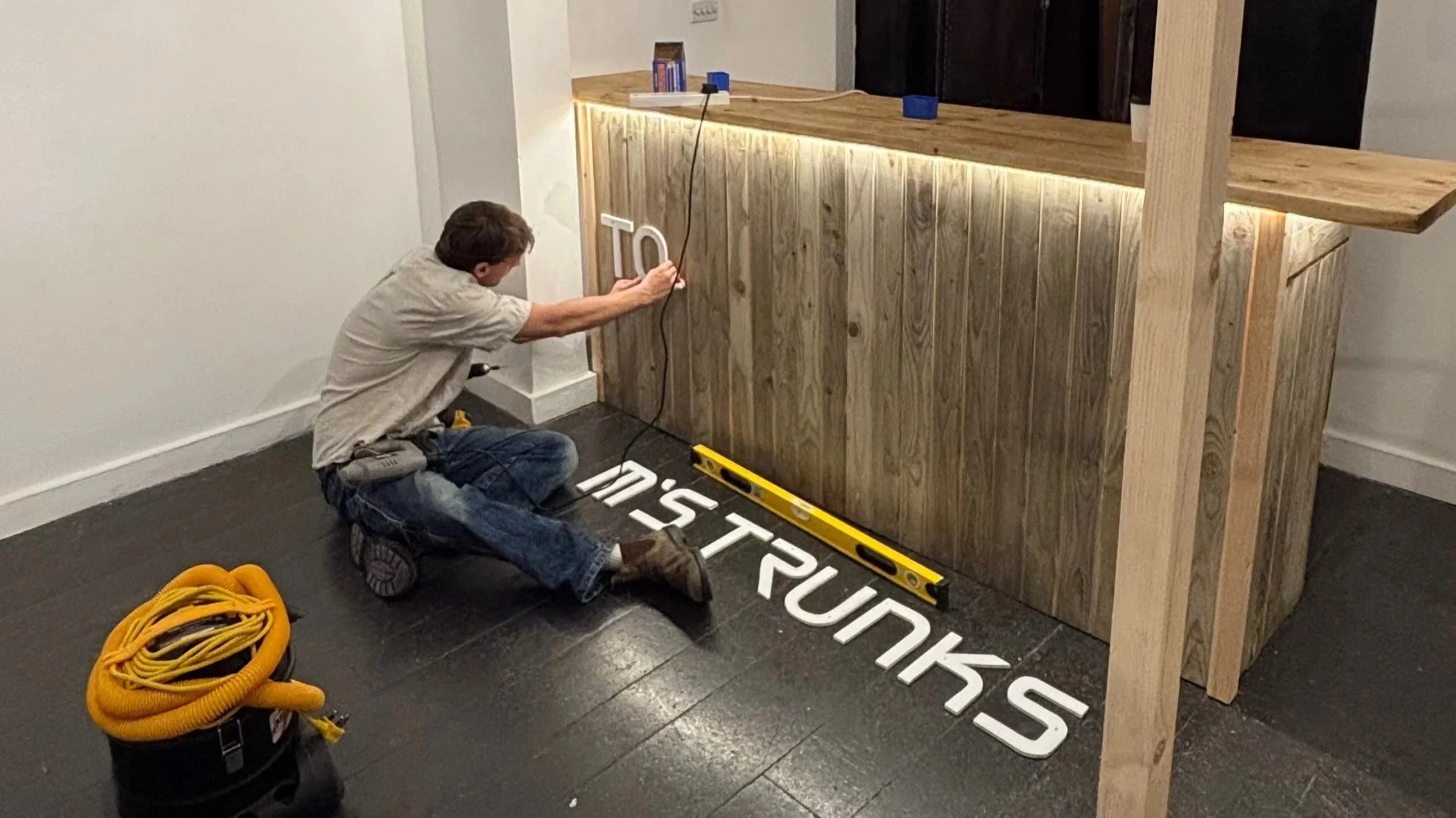 A person kneeling on the floor assembling a wooden bar counter. They are attaching a white letter sign to the counter. The person is wearing a beige shirt and jeans, with various tools and supplies nearby, including a yellow extension cord, a level, 