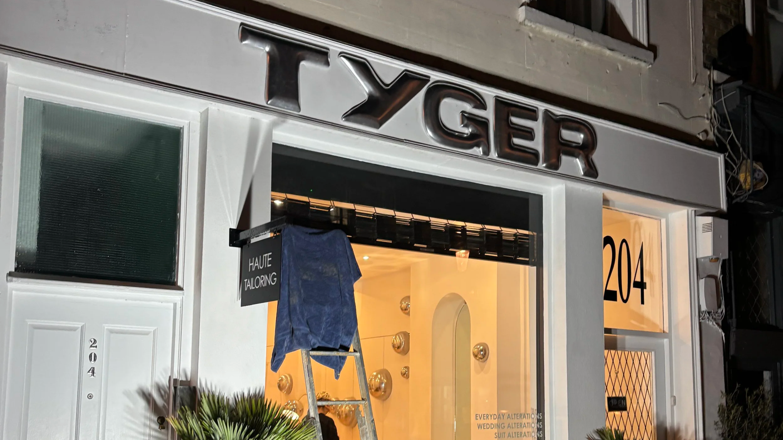 Storefront with illuminated sign reading 'TYGER', window display with decorative gold spheres and mannequin, address 204, and a sign indicating Haute Tailoring and wedding alterations.