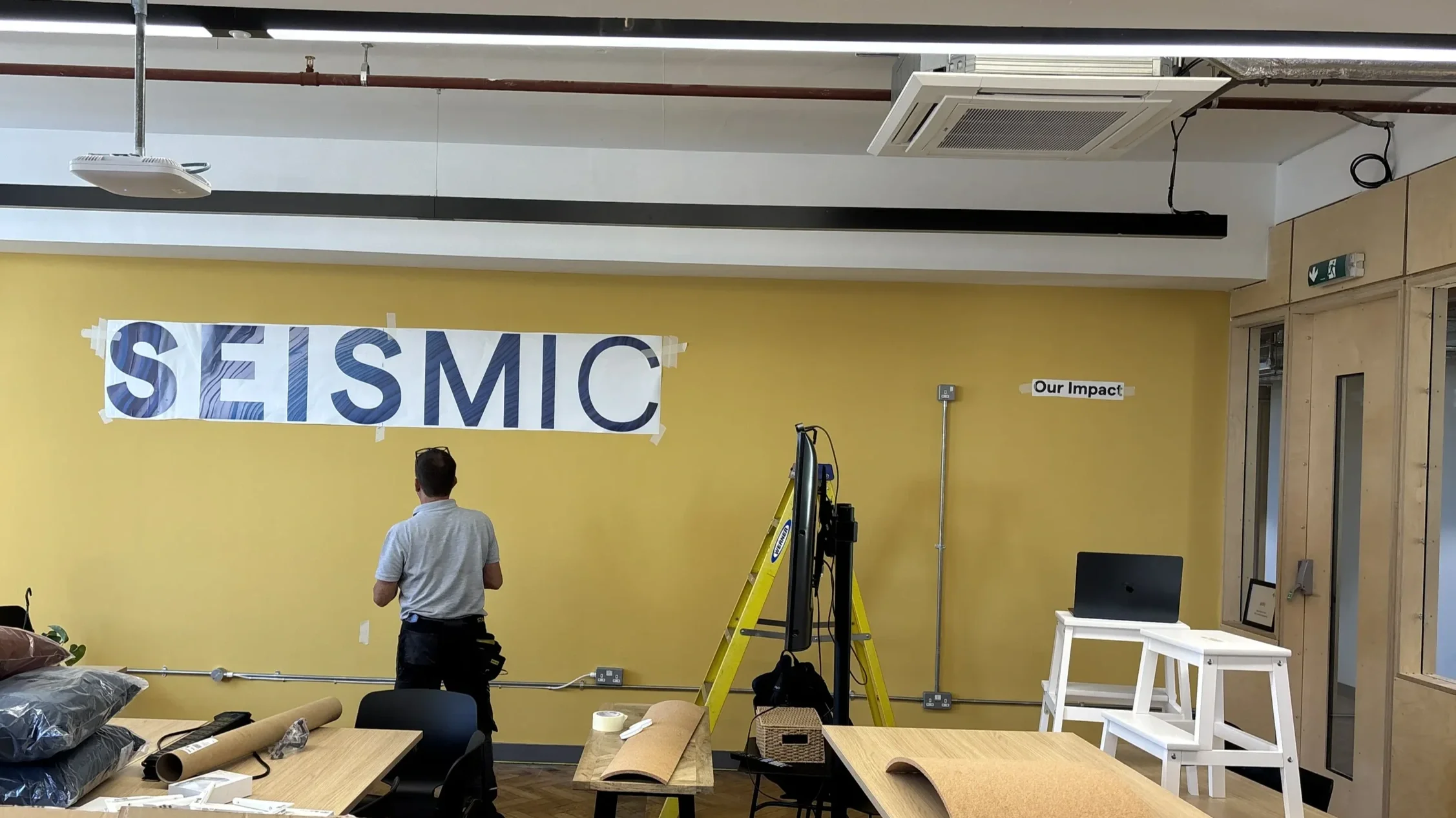 Seismic office brand installation in progress with two workers fitting large lettering on yellow feature wall using a ladder