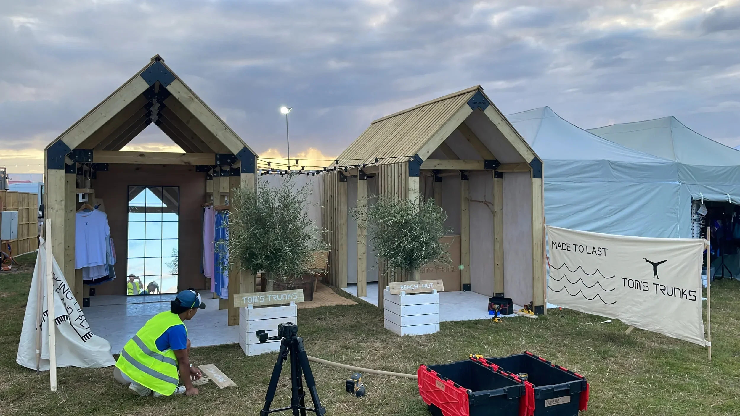 Tom's Trunks bespoke wooden beach hut brand activation structures being set up at outdoor festival with worker on site