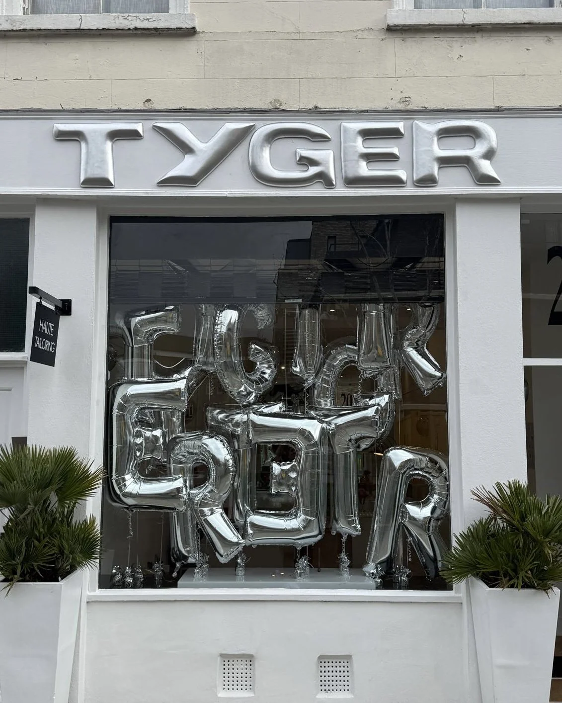 Storefront with metallic lettering and balloons spelling out "FESTIVE," with the store name "TYGER" above, and potted plants outside.