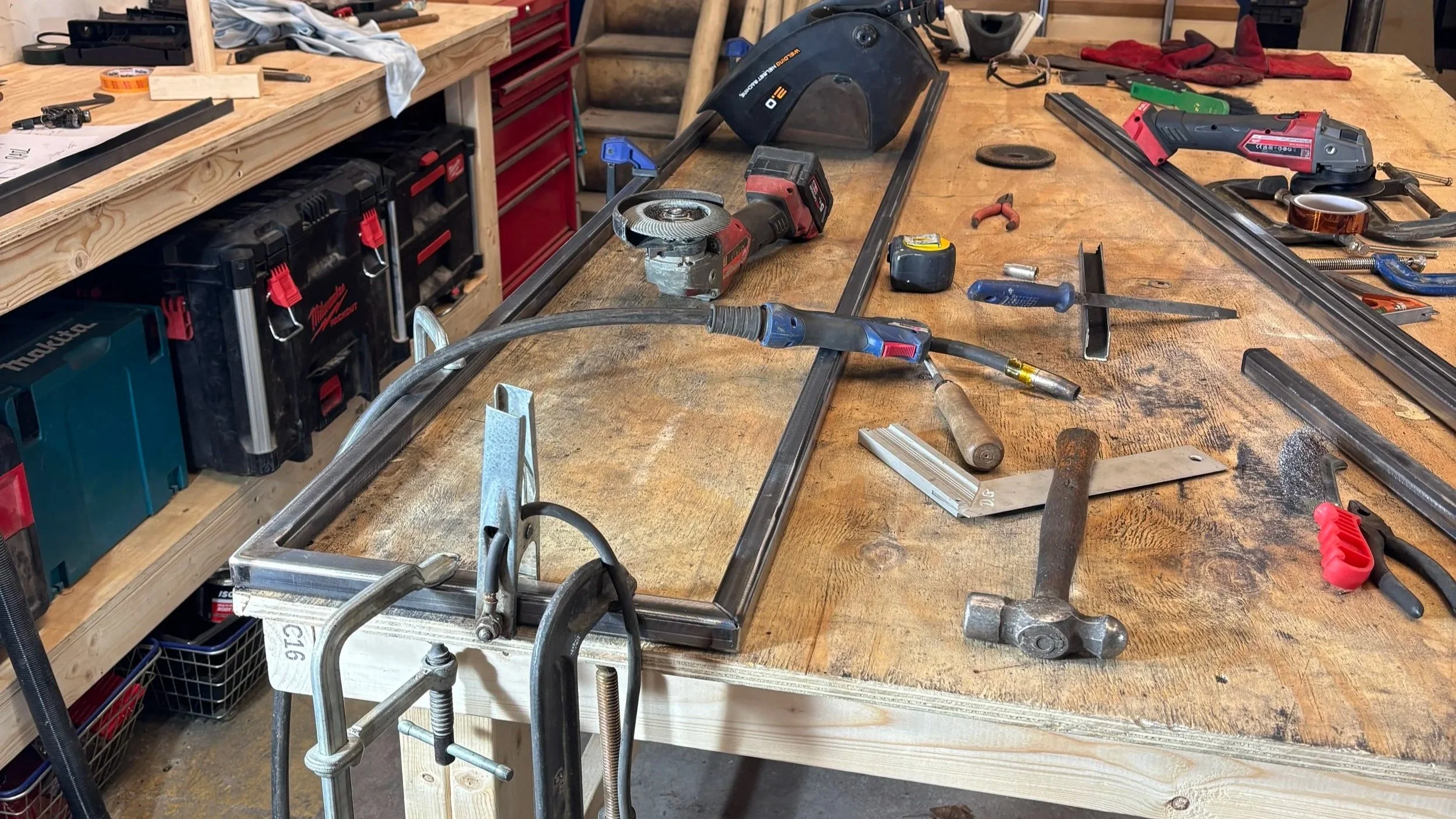 Workshop workbench with various tools, including a circular saw, a cordless drill, a hacksaw, a clamp, and measuring tools. The surface is cluttered with metal tracks and accessories. In the background, there are red tool chest drawers and shelves with additional tools and materials.